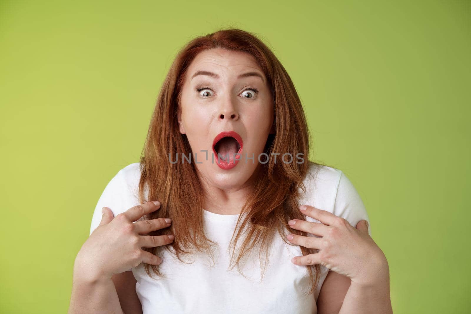 Shocked panicking redhead middle-aged woman gasping drop jaw open mouth stare camera freak-out anxious pointing herself impressed terrified frustrated nervously react green background.