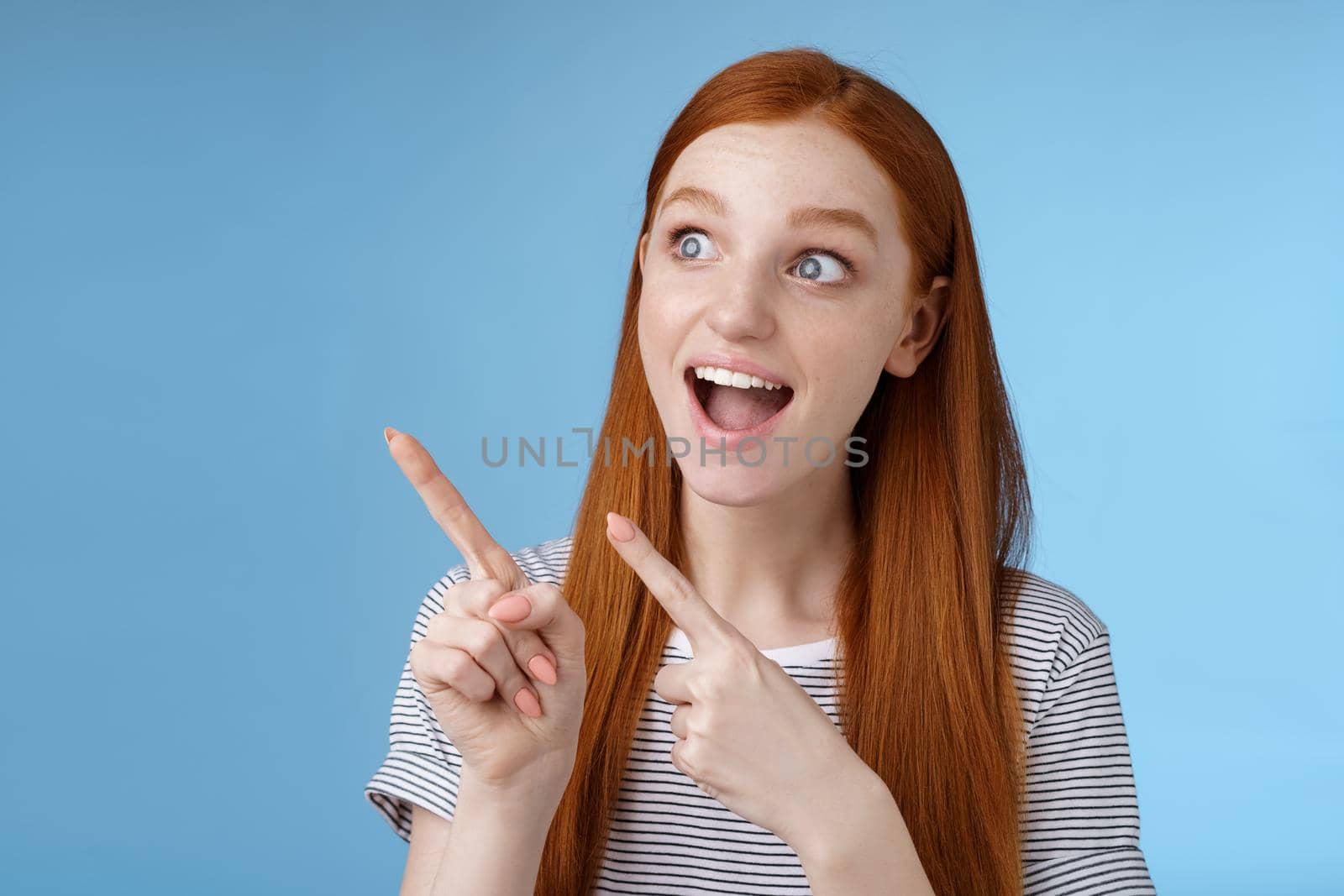 Amused adorable redhead european girl say wow stare gasping astonished pointing looking upper left corner excited smiling drop jaw amazed standing thrilled blue background excitement concept by Benzoix