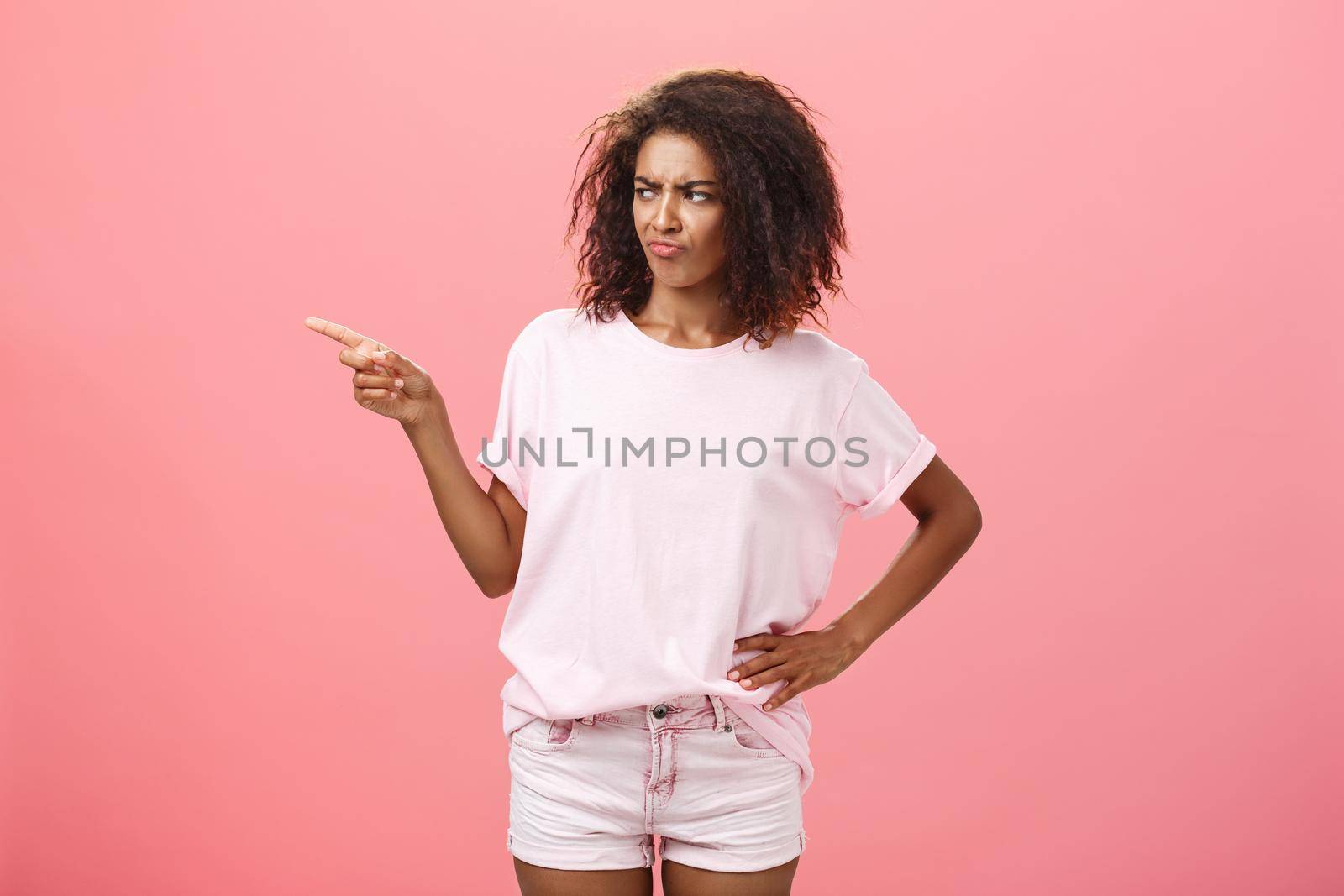 Picky fashionable and attractive young female stylist with curly hairstyle frowning and pouting with dislike and doubt looking and pointing left unsure having hesitation look fits customer. Body language and advertisement concept