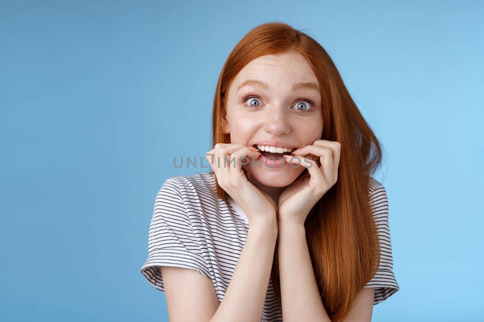 Excited amazed attractive redhead girl blue eyes look fascinated affection smiling desire bite fingernails eager feel astonished cannot wait bite tasty food, standing blue background thrilled by Benzoix