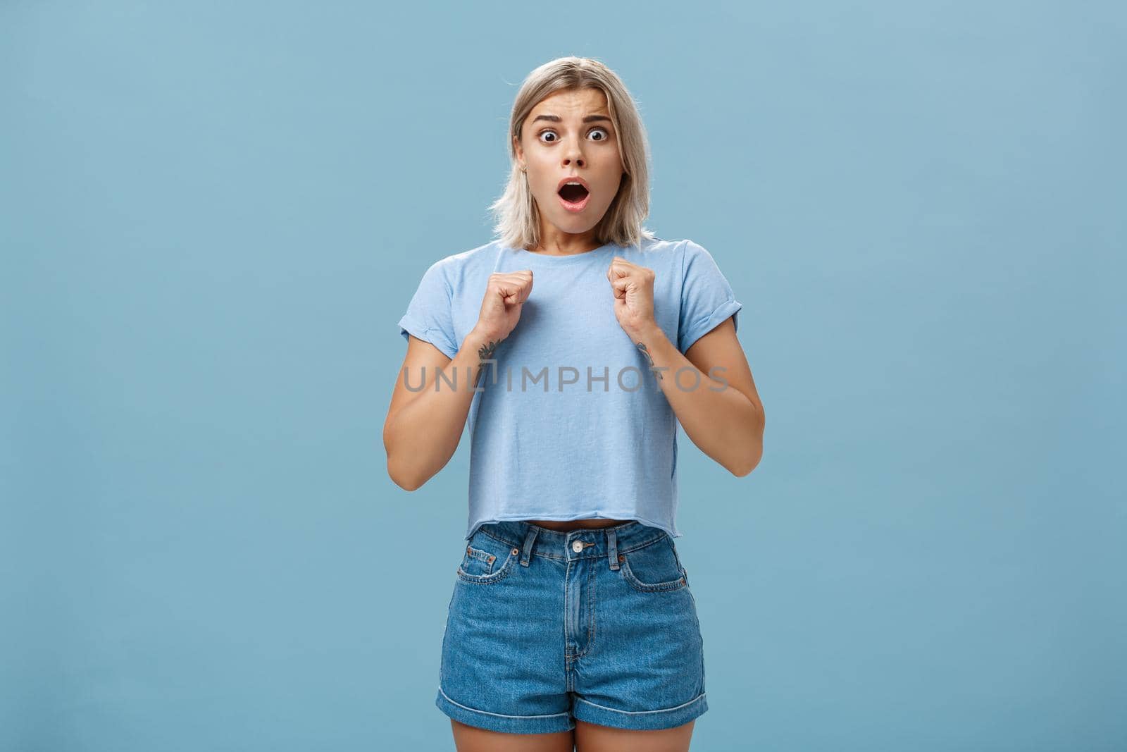 Studio shot of stunned shocked girl standing in stupor with dropped jaw and frightened look clenchign fists near breast from fear standing astonished over blue background. Emotions concept