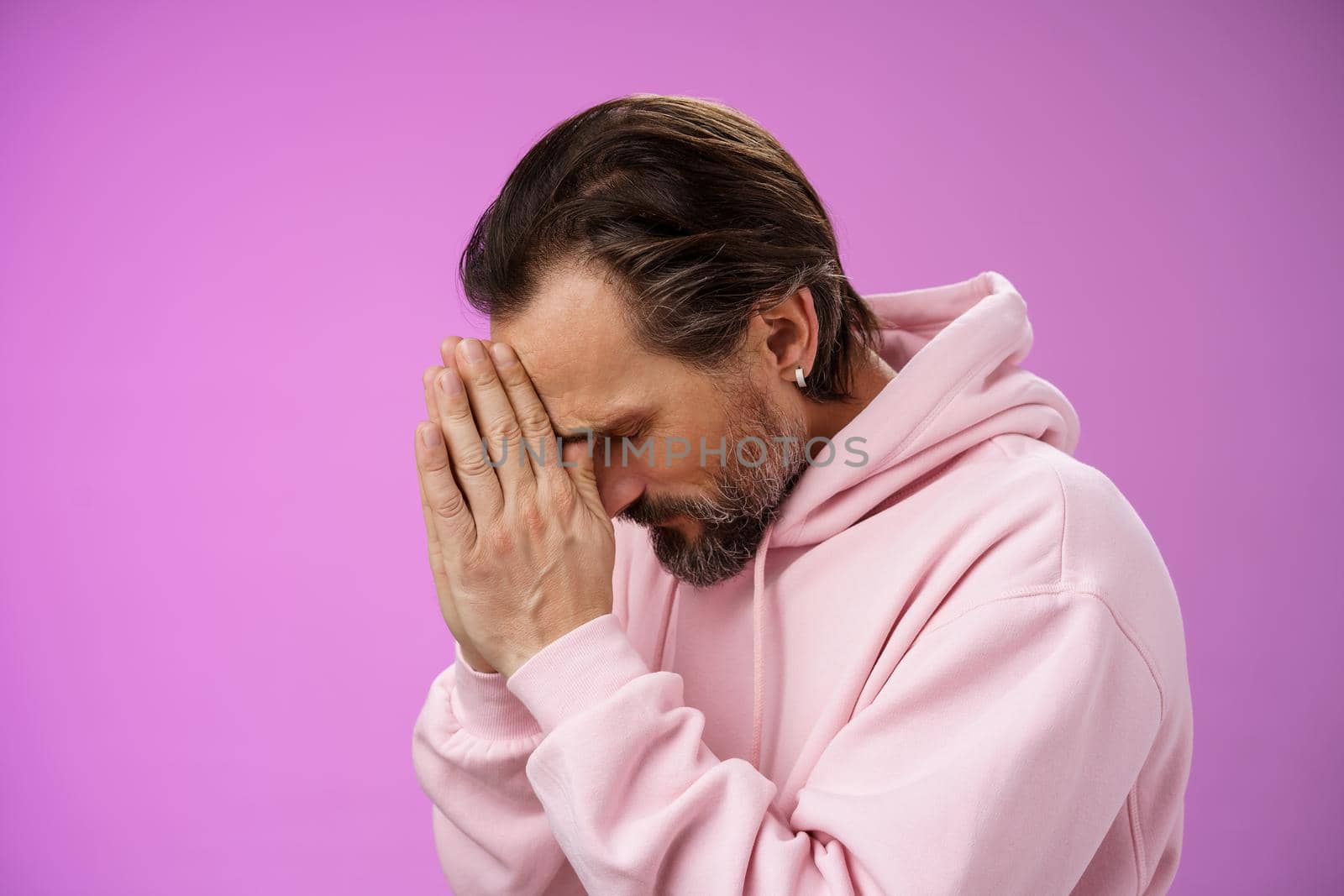 Concerned upset man supplicating asking god mercy help praying bow head close eyes hold hands pray hopefully waiting miracle worried wife health, standing sadness purple background. Copy space