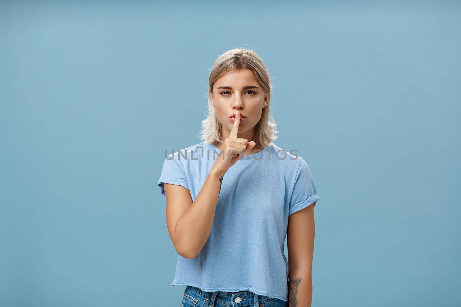 Promise not tell anyone it secret. Mysterious attractive and feminine blond woman with tanned skin and tattoos shushing with index finger over mouth squinting as if hiding something over blue wall.