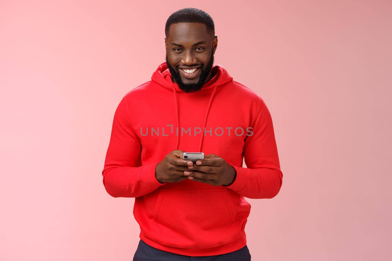 Attractive happy smiling african american bearded guy in red hoodie playing games smartphone grinning joyfully messaging having interesting online chat, standing satisfied pink background.