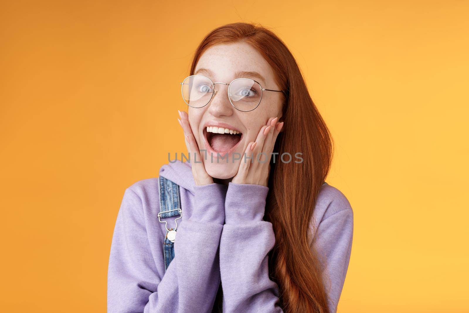 Excited happy smiling broadly redhead girlfriend drop jaw grinning thrilled look left astonished touch cheeks reacting awesome stunning news screaming happiness, standing orange background.