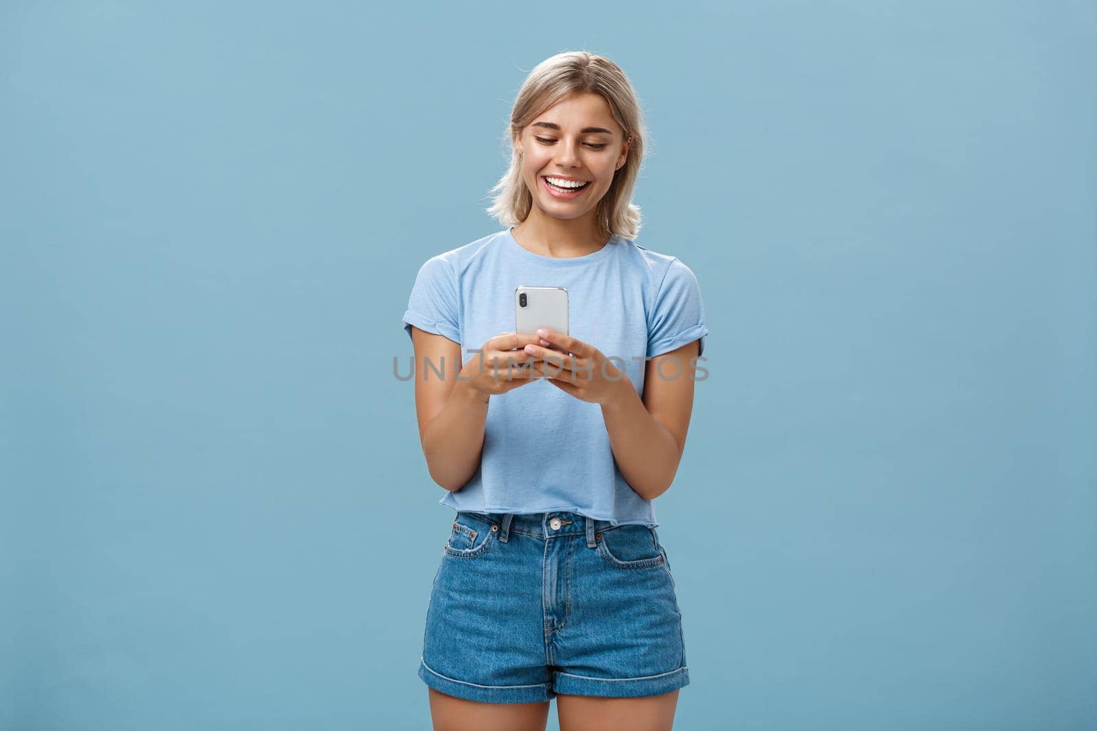 Nice to have internet friends. Carefree happy attractive blonde female student with tanned skin and blond hair reading message from smartphone smiling at device screen pleased over blue wall by Benzoix