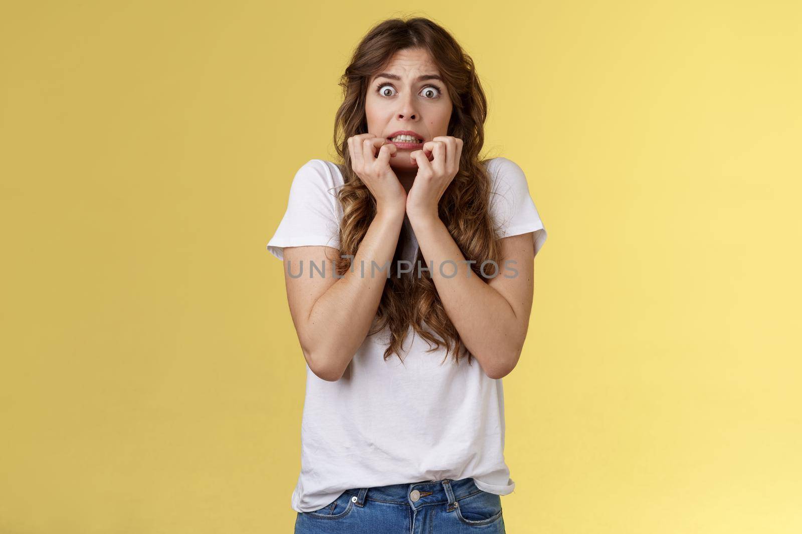 Scared timid insecure unconfident european girl curly haircut hold hands face biting fingers look frightened shocked standing stupor speechless shaking fear terrified yellow background by Benzoix