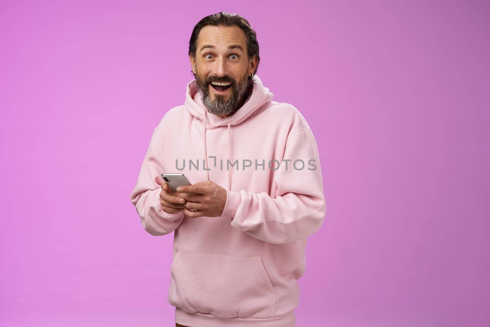 Amused happy fascinated adult mature caucasian bearded man in pink hoodie gasping impressed holding smartphone look camera astonished purchase cool new phone, astonished how convenient.