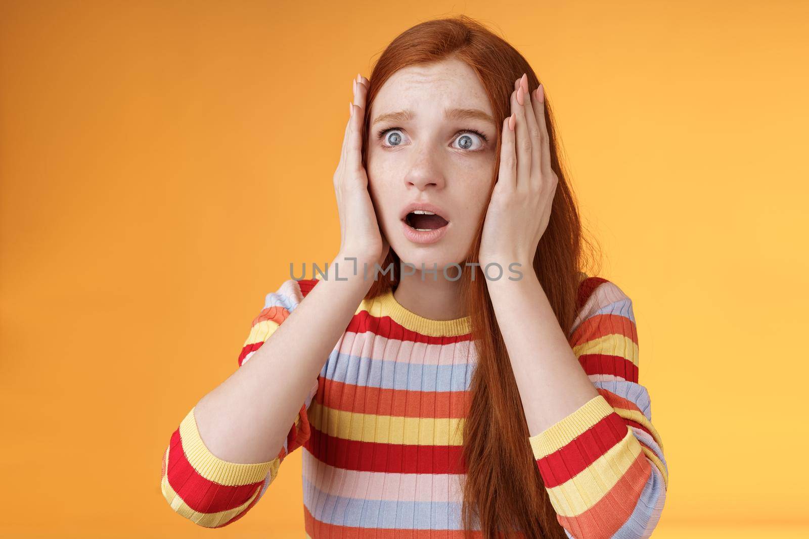 Worry insecure redhead european panicking girl grab head hands both sides gasping open mouth shocked stare frightened upset watching terrible accident standing concerned orange background.