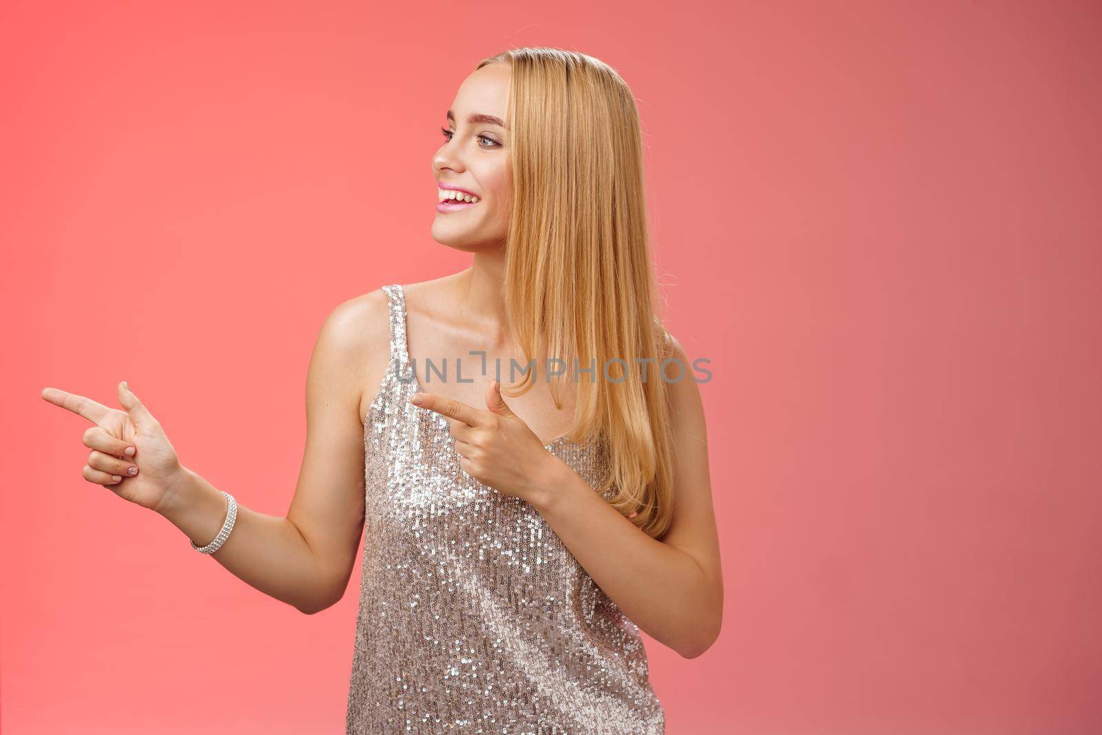 Joyful carefree young wealthy glamour blond woman in glittering silver dress enjoying awesome party dancing turning pointing left smiling broadly waiting girlfriend bring drinks, red background.