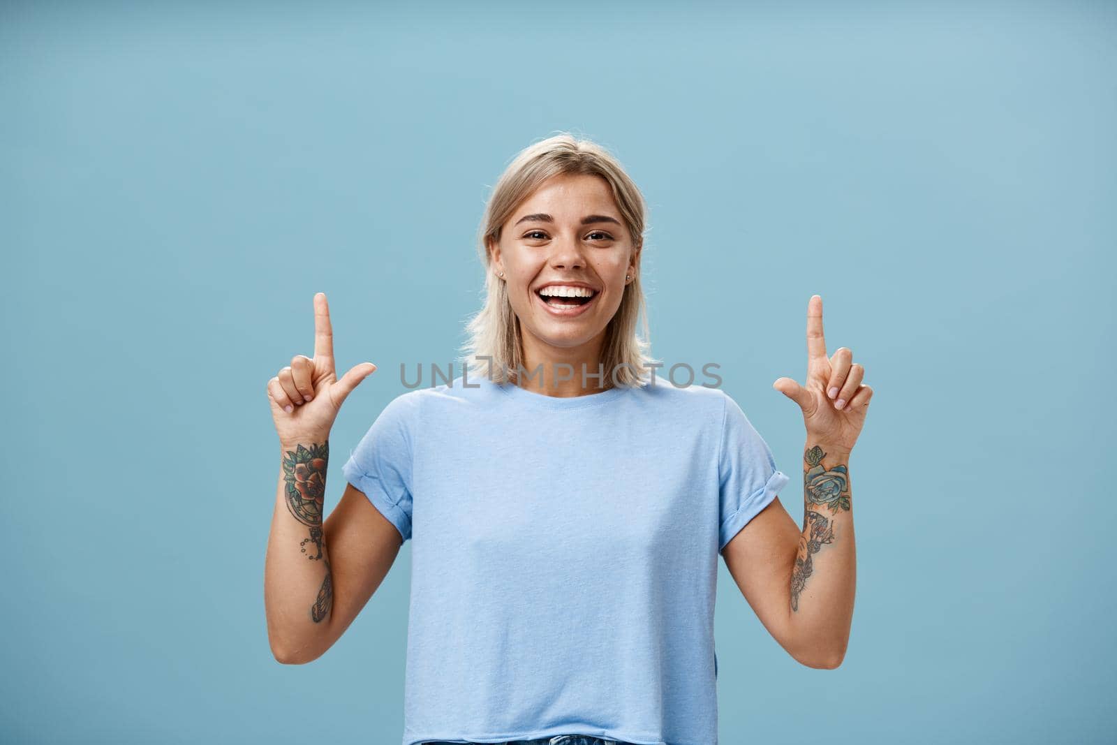 Waist-up shot of upbeat creative stylish and gorgeous blonde female student have tattoos on arms pointing up with raised forefingers smiling with delight and joy posing over blue background. Advertisement concept
