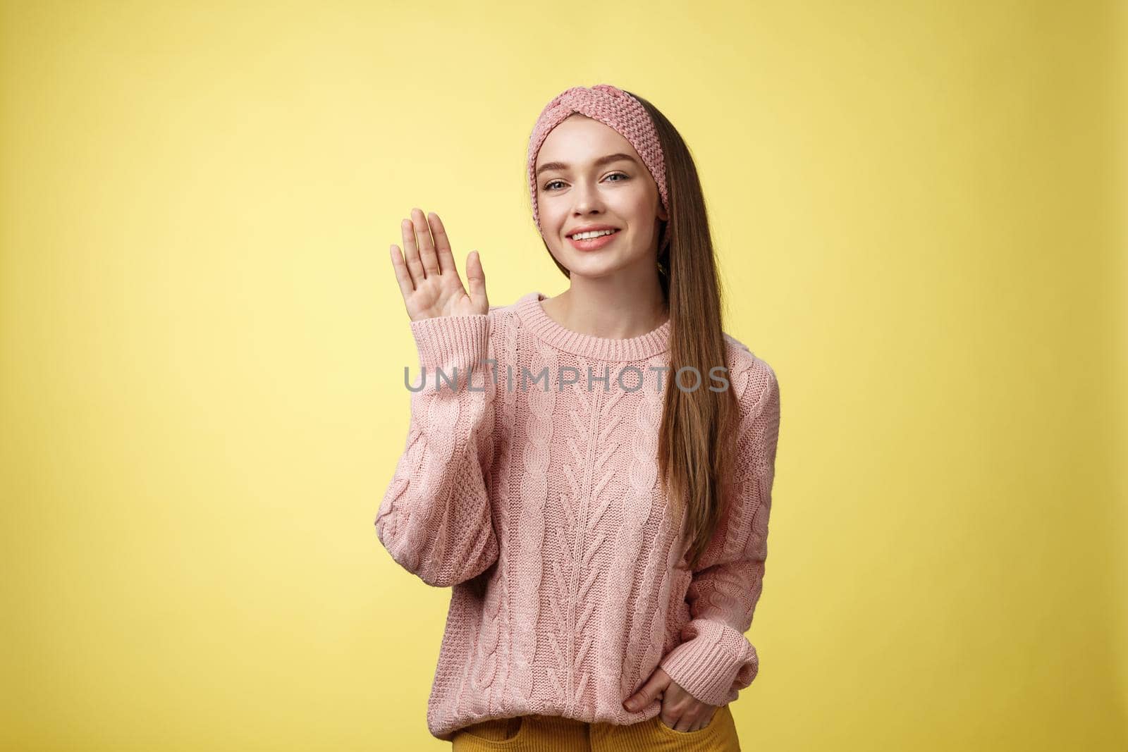 Hello adventures. Charming glamour 20s girl in sweater, knitted headband waving palm saying hi friendly smiling, holding hand in pocket gretting friends, posing positive and carefree over yellow wall.