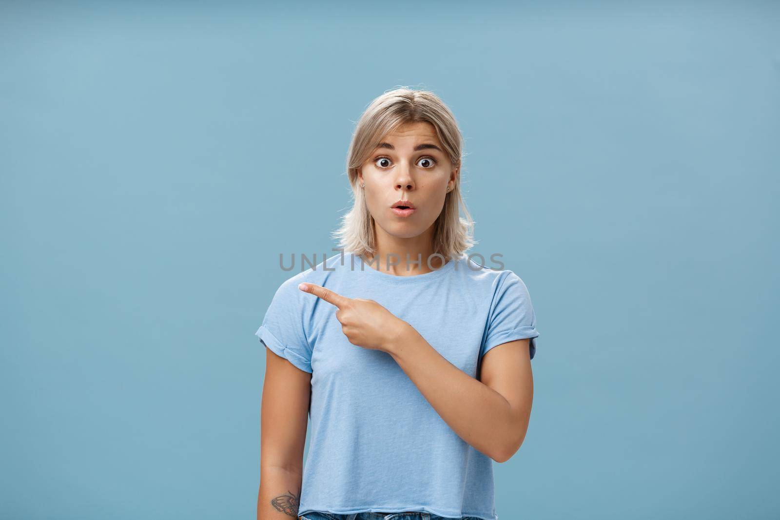 Studio shot of amazed stunned cute blonde witnessing unbelievable event gasping opening mouth staring astonished and pointing left being questioned and shocked over blue background. Emotions concept