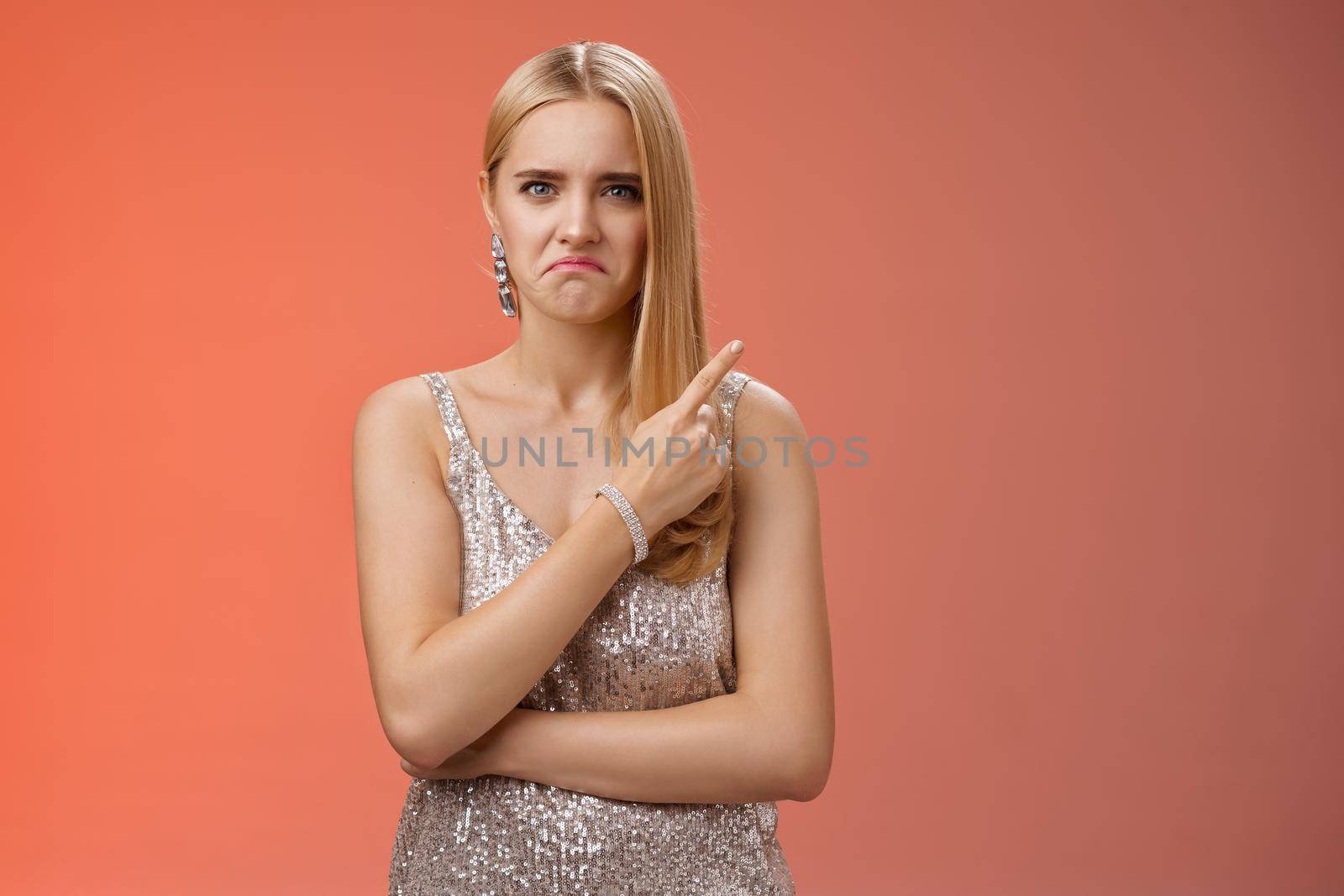 Jealous displeased angry young revengeful blond ex-girlfriend in luxurious silver shiny dress frowning pouting pointing upper right corner displeased pissed standing fed up upset red background.