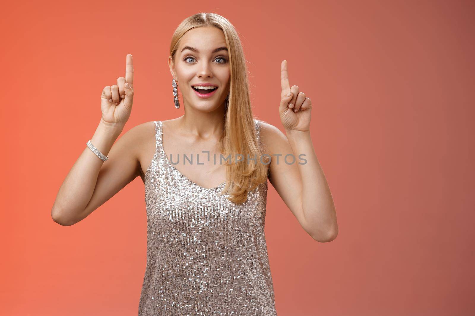 Charming amazed blond european woman in fabulous silver glittering dress raise hands point up amused enjoying watching shooting stars, fireworks gaze camera excited happy surprised, red background.