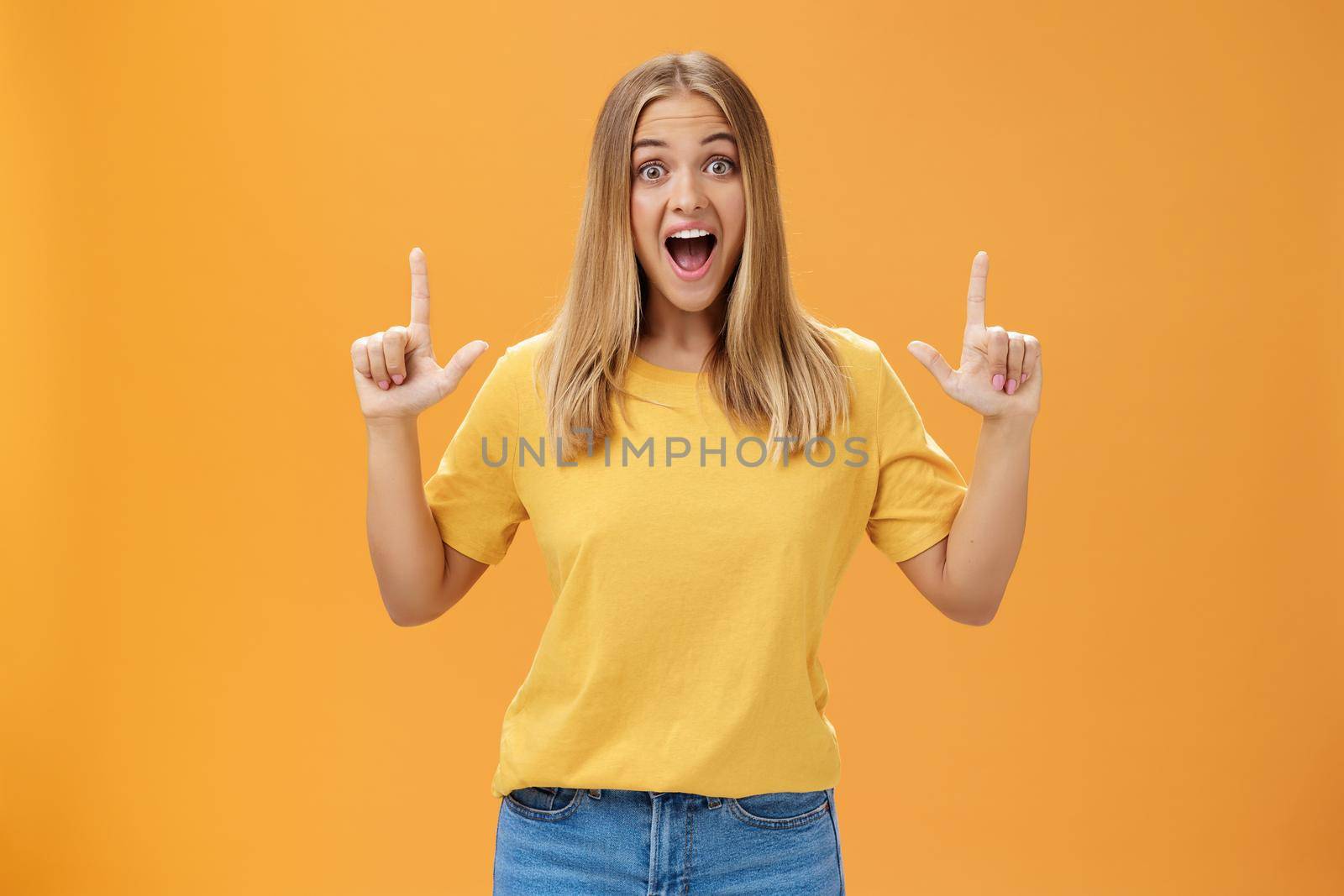 Portrait of joyful surprised and impressed delighted young woman with tanned skin and fair hair opening mouth from amazement and joy raising hands pointing up at awesome copy space against orange wall by Benzoix