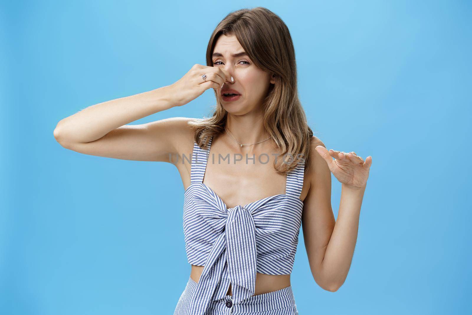 Gross smells awful. Portrait of displeased funny young slim woman with tattoo squeezing nose with two fingers not sniff disfusting reek crying from stink standing intense over blue background.