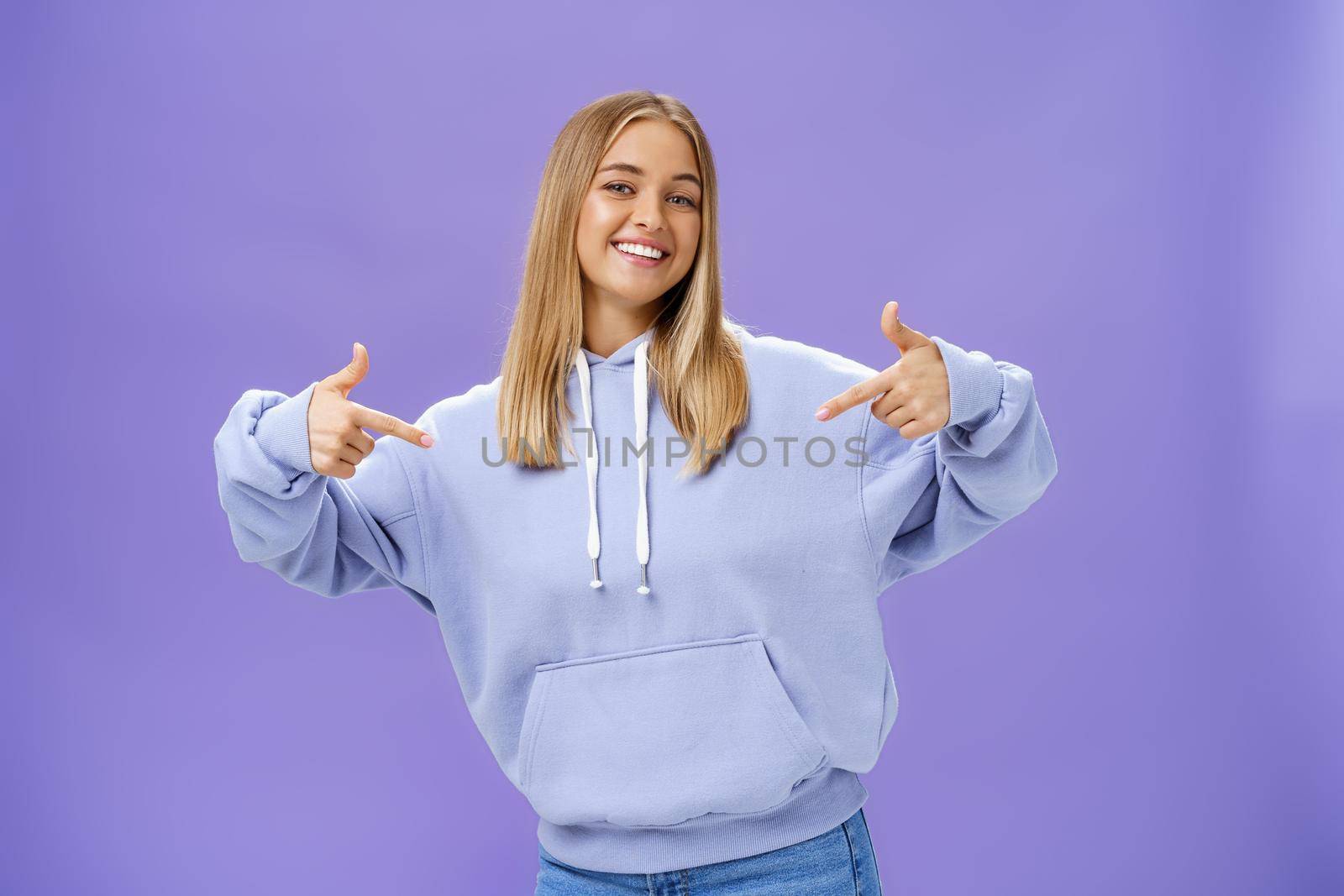 Self-assured cheerful carefree young girl with tanned skin and fair hair pointing proudly at herself bragging about own skills and achievements standing cool feeling awesome in new hoodie. Body language and attitude concept