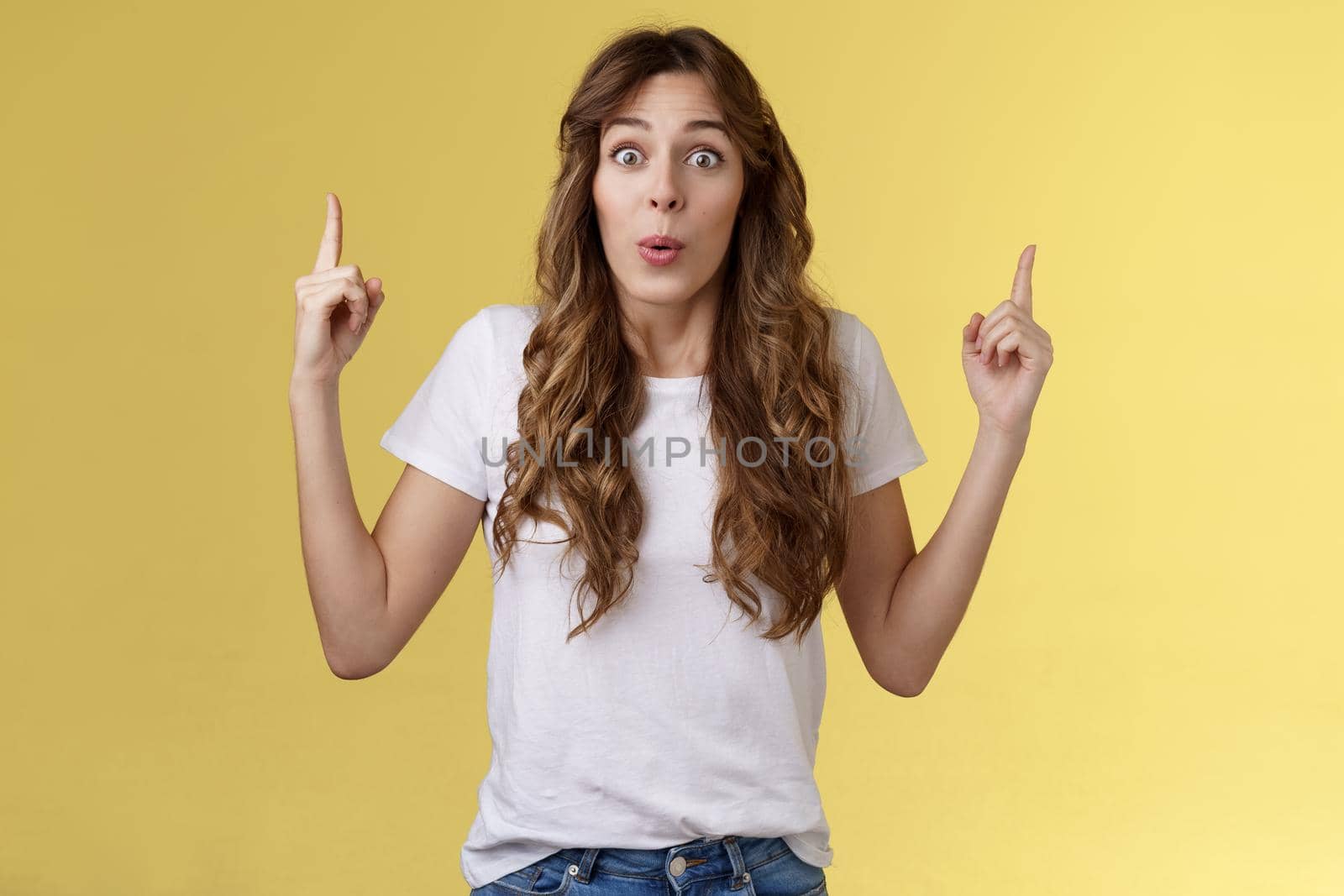 Impressed enthusiastic surprised ambushed cute female model fold lips wow admiration joy stare camera fascinated pointing up index fingers top copy space show you stunning offer yellow background. Lifestyle.