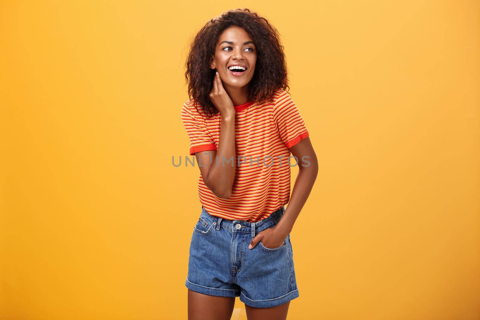 Stylish feminine and fashionable african american female model with afro hairstyle touching neck gently looking right with amused carefree expression holding hand in pocket over orange background. Lifestyle.