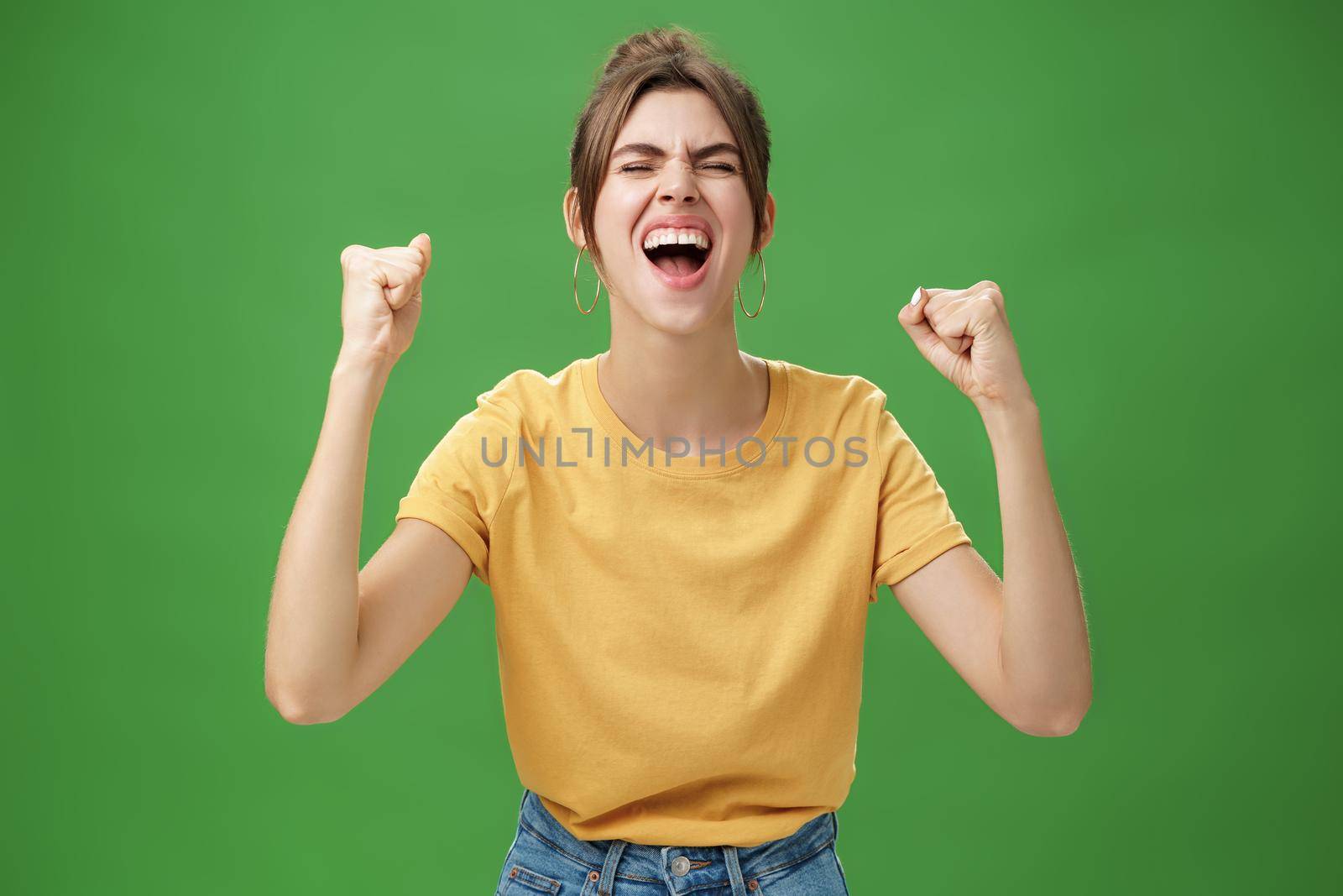 Waist-up shot of charismatic energized and excited female in yellow t-shirt closing eyes yelling from joy and happiness raising hands in cheer celebrating successful news over green background. Lifestyle.
