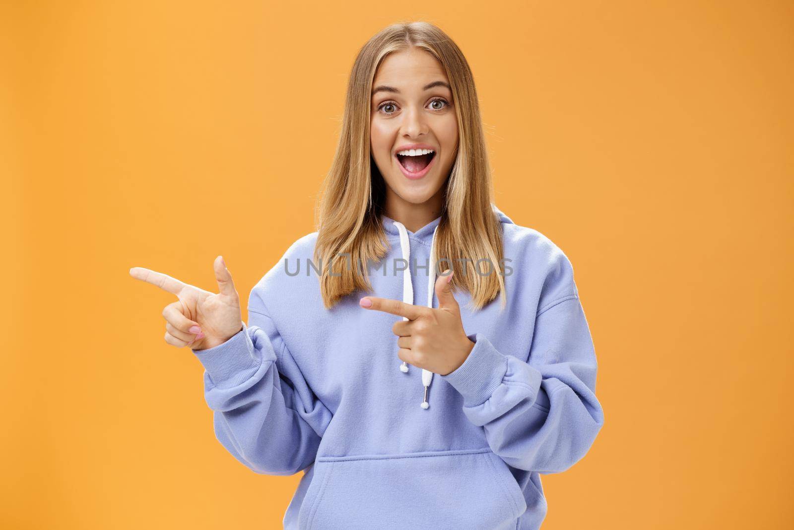 Amused girl pointing at curious copy space showing left with index fingers smiling excited and surprised standing entertained with upbeat grin in cozy blue hoodie over orange background by Benzoix