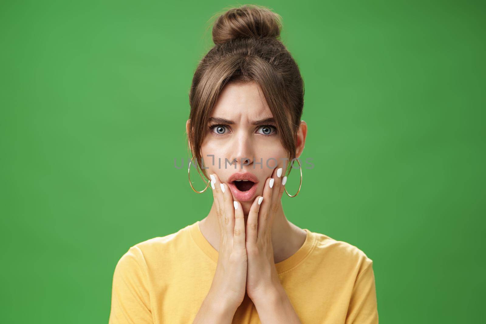 Shocked woman reacting to pimple looking disturbed and displeased in mirror holding hands pressed to cheeks open mouth and frowning being disappointed and upset over green background by Benzoix