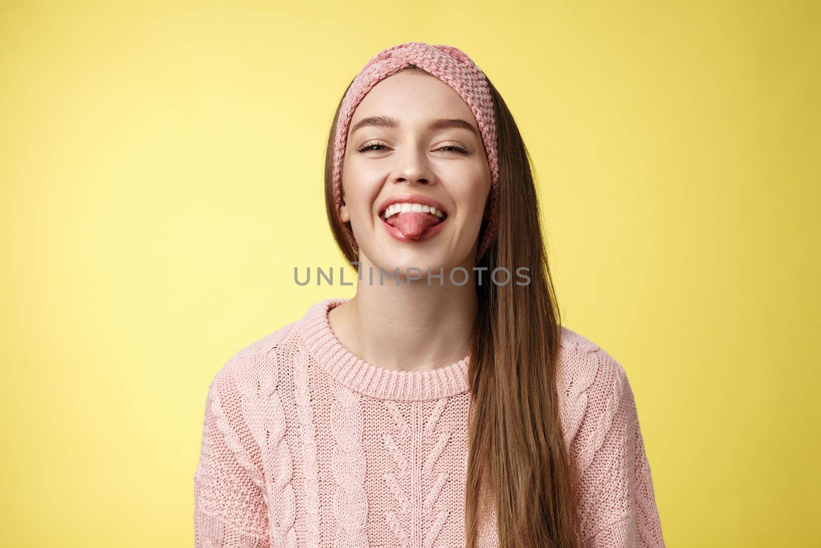 Positive entertained cute funny glamour young european girl in sweater, knitted trendy headband smiling fooling around showing tongue playfully, mocking friend enjoying sunny day over yellow wall.