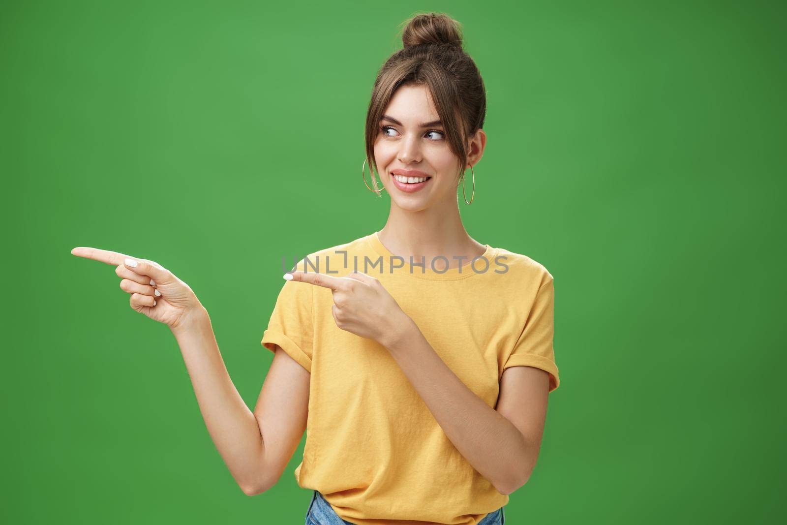 Waist-up shot of good-looking friendly and feminine female coworker in yellow t-shirt showing newbie office pointing and looking left with cute happy smile posing against green background. Advertisement concept