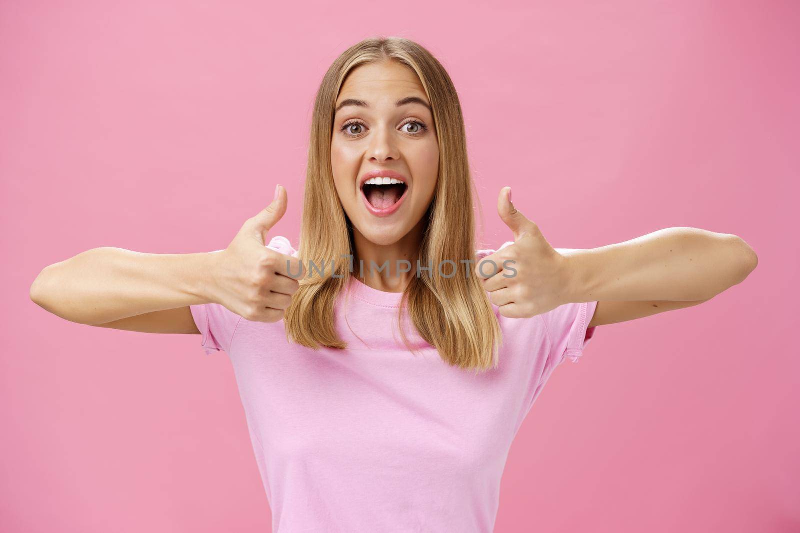 Supportive girlfriend cheering for favorite player showing thumbs up with happy positive smile giving approval and showing she likes perfect idea posing amused and enthusiastic against pink background by Benzoix