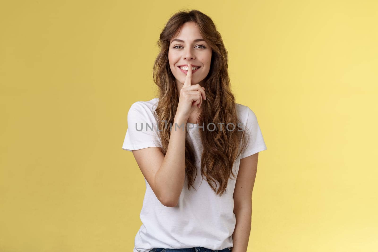 Cute cunning lovely european girl curly hairstyle hiding beauty secret smiling sensually show hush shush gesture index finger pressed lips grinning joyfully stand yellow background by Benzoix