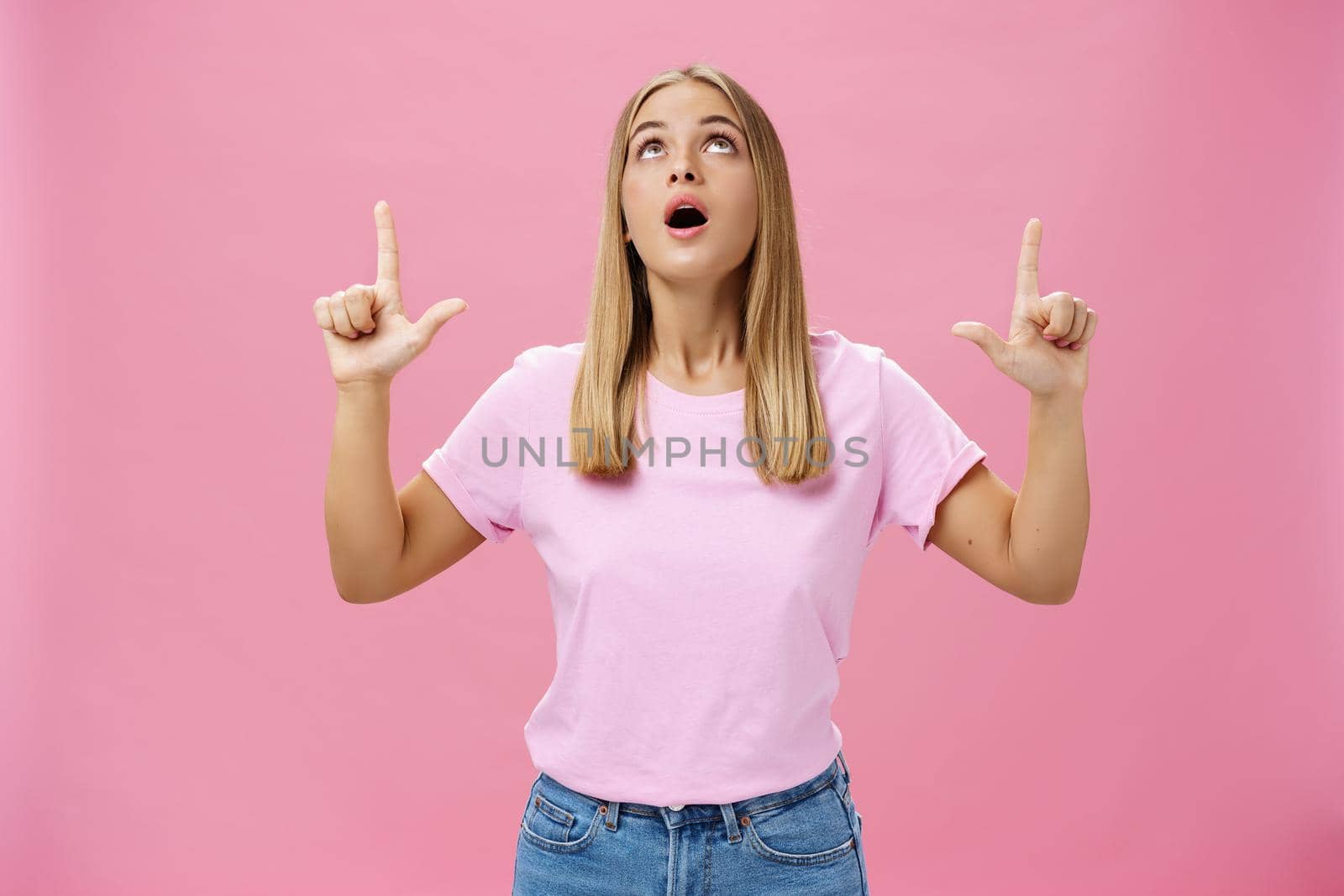 Portrait of amused and impressed curious attractive woman in t-shirt dropping jaw looking and pointing up astonished and intrigued watching interesting object in sky posing against pink background. Lifestyle.