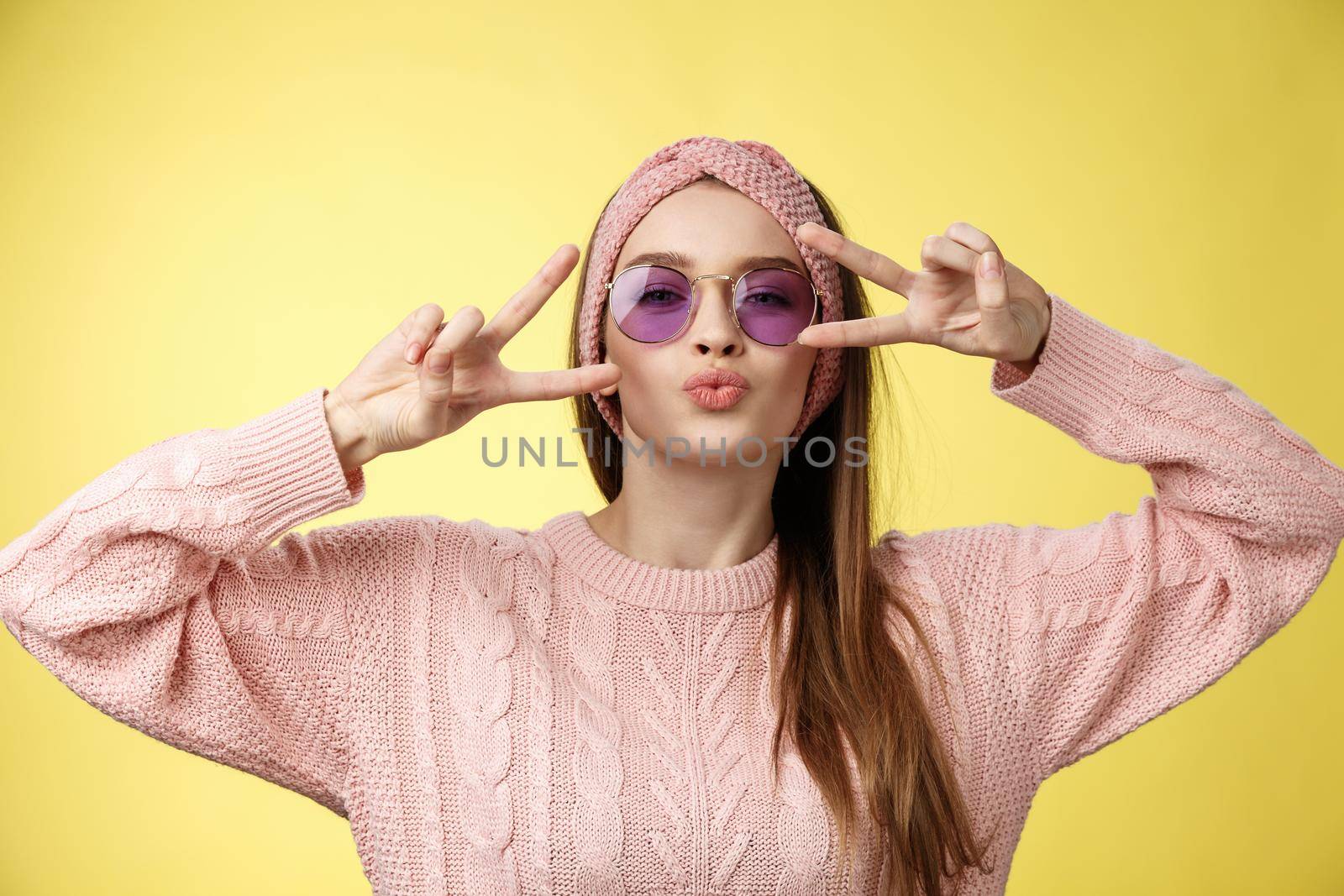Mwah followers. Cheeky and sassy cute glamour young european girl wearing sunglasses, sweater showing disco gesture peace, victory over eyes folding lips, blowing kiss at camera against yellow wall.