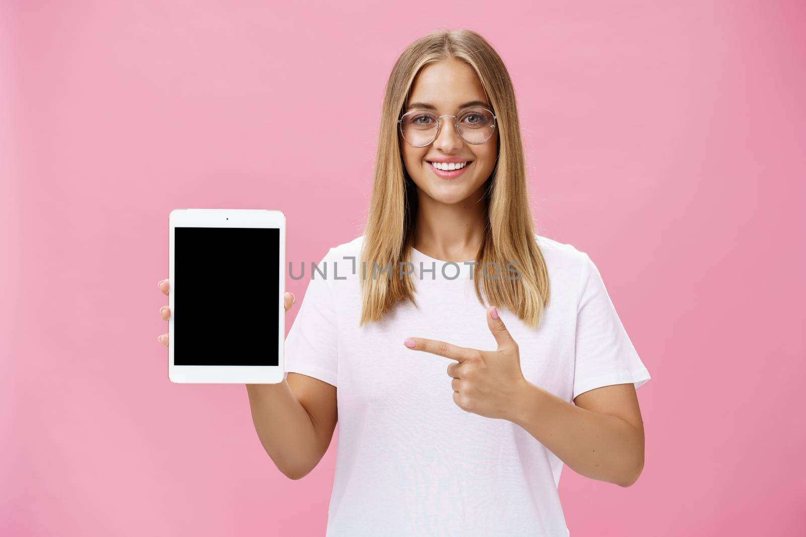 Waist-up shot of optimistic and joyful female showing cool digital tablet pointing at device screen and smiling broadly at camera giving advice what app useful posing in glasses against pink wall. Lifestyle.