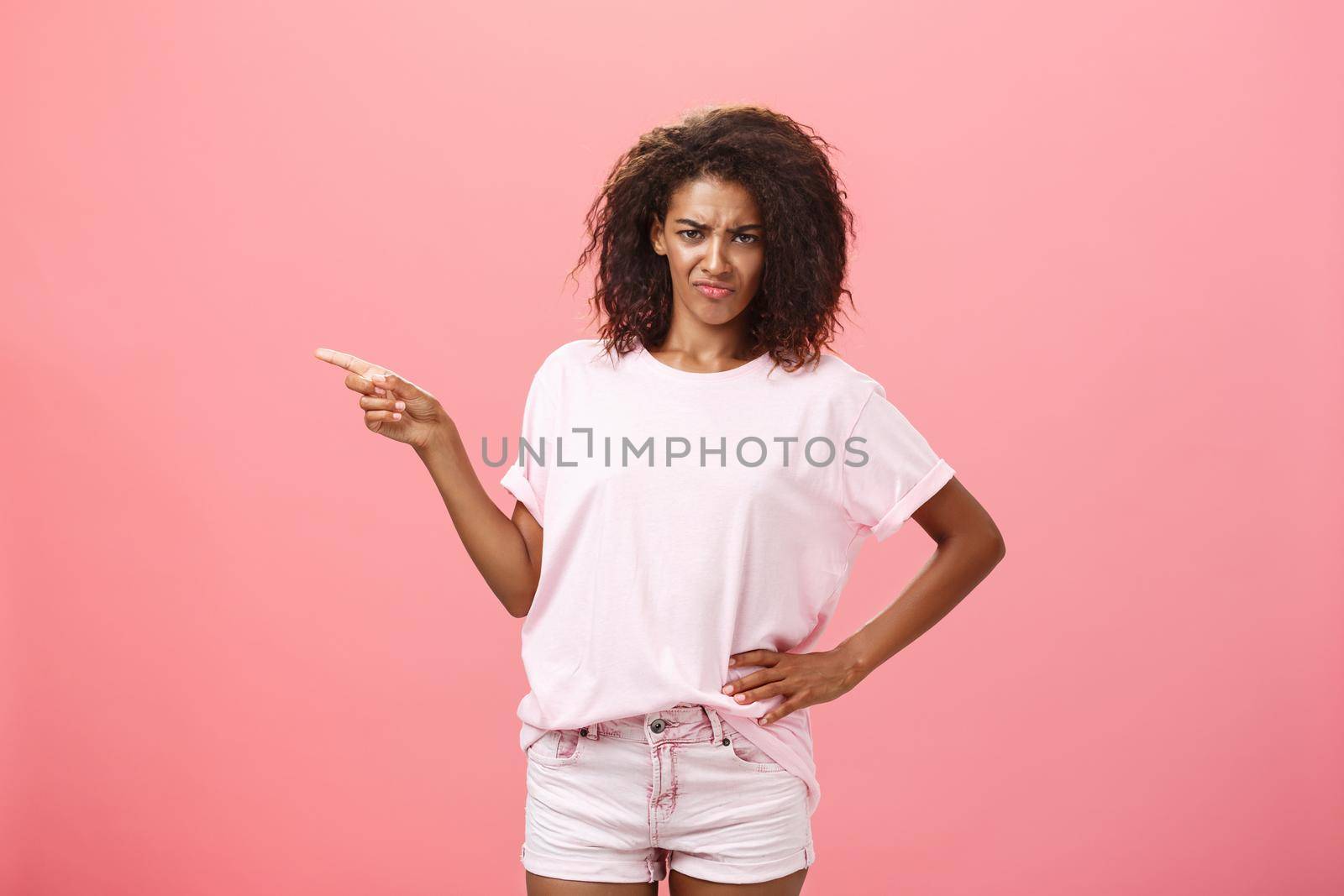 Girl feeling offended seeing dissatisfying product. Portrait of displeased serious-looking bothered dark-skinned female in t-shirt frowning from dislike pointing at creepy thing over pink background.