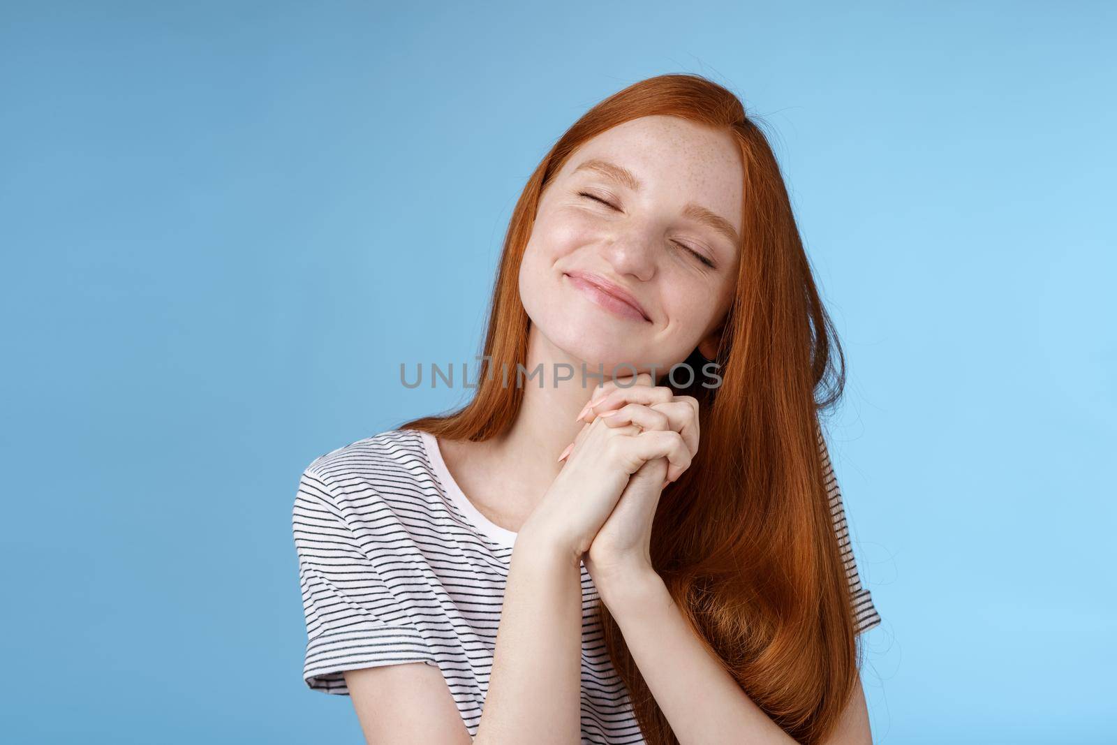 Charming delighted happy dreamy redhead girlfriend ginger long hair tilting head close eyes smiling pleased daydreaming thinking about sweet tender memories press palms together, blue background.