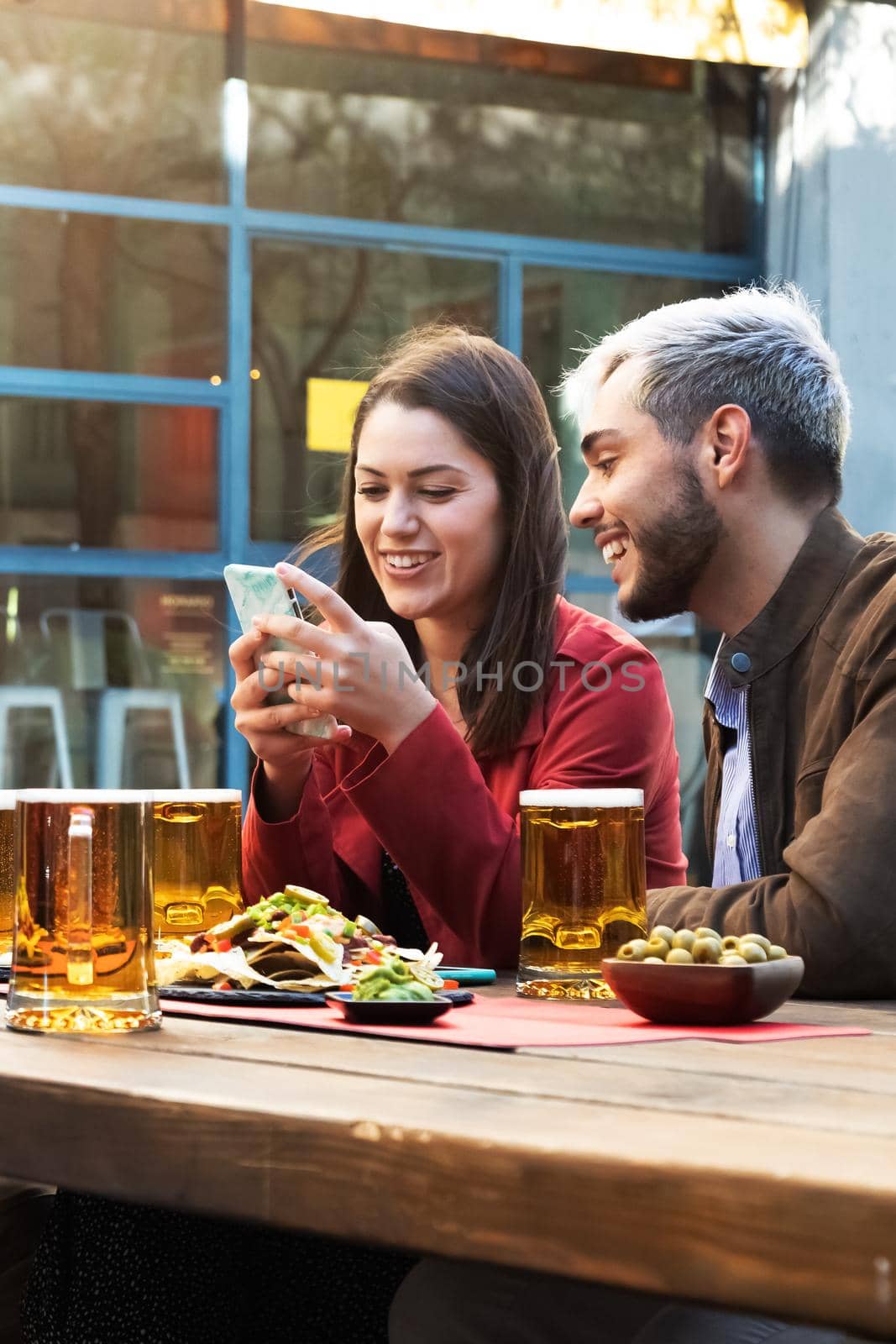 Friends looking at mobile phone in a bar enjoying beer and appetisers. Vertical image. Friendship concept.
