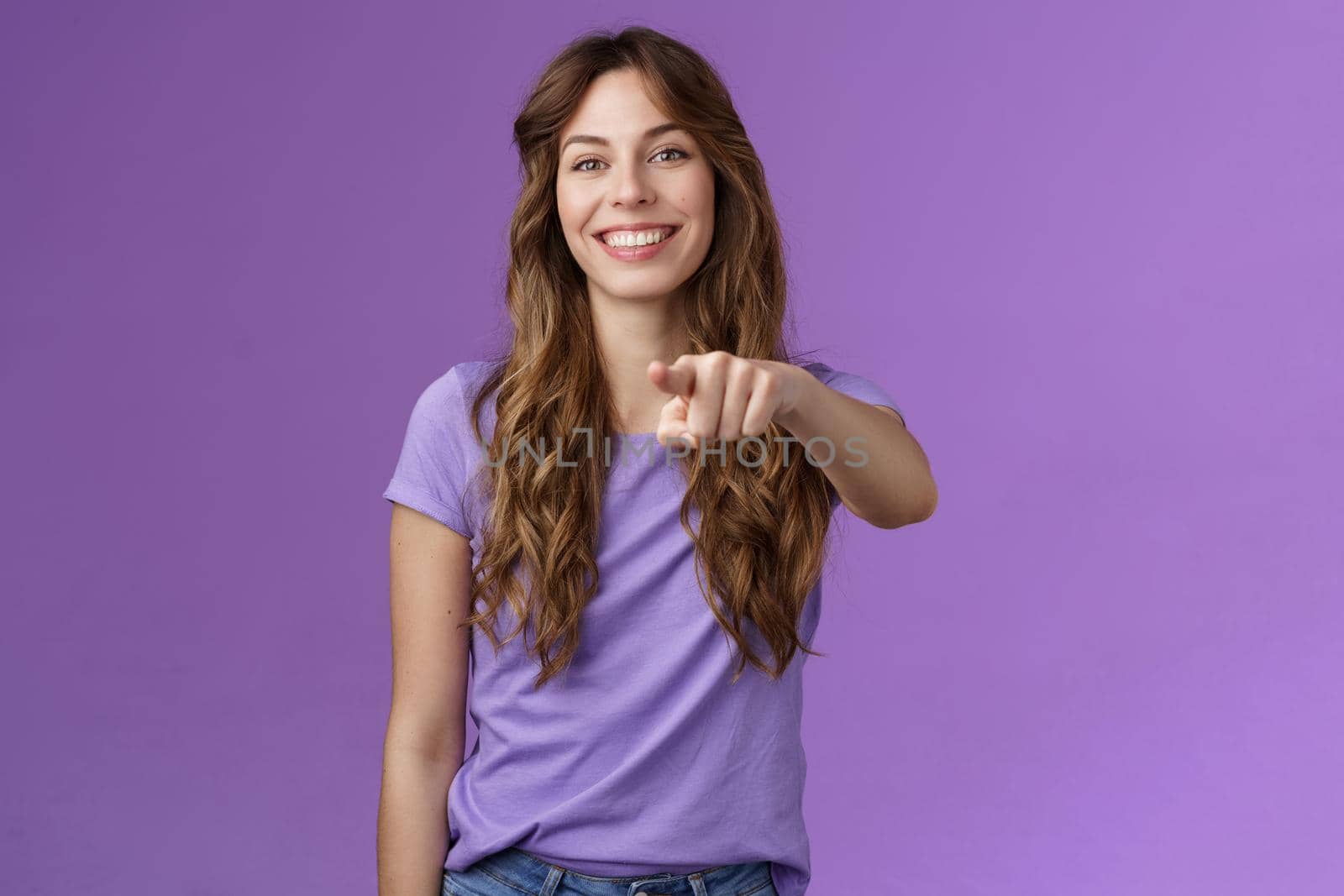 Cheerful girl pointing you. Attractive friendly happy smiling curly-haired female deciding who pick indicating finger camera grinning joyful inviting coworker her team stand purple background.