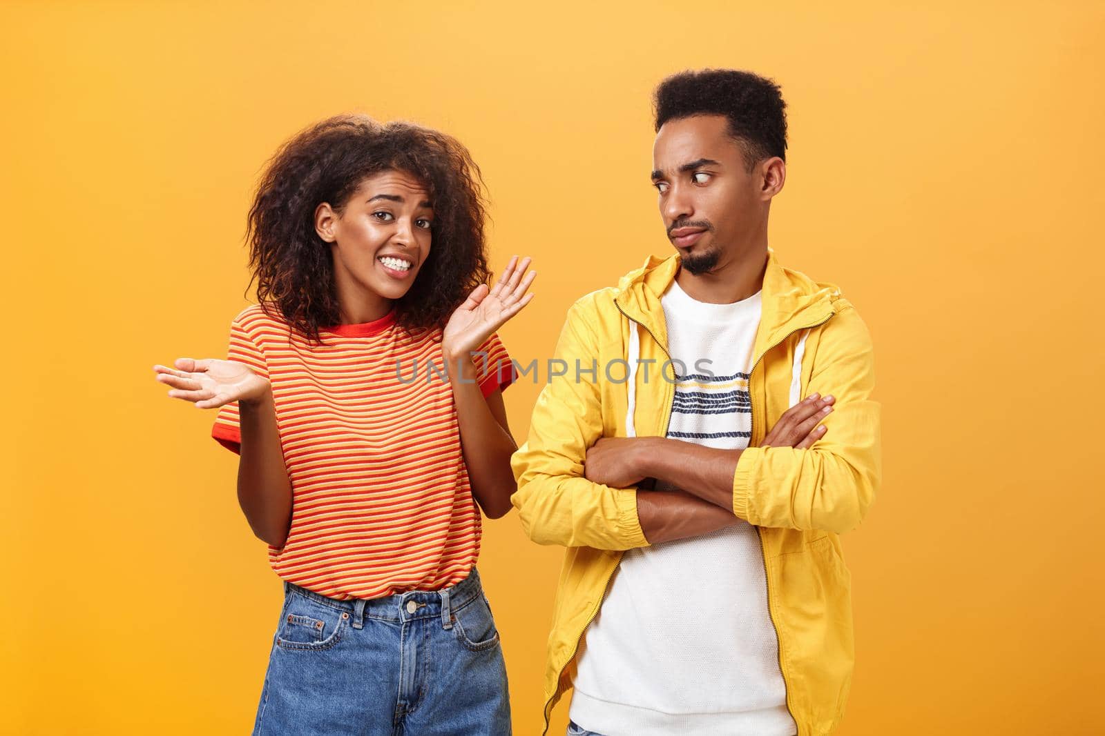 Guy thinks his friend weirdo making dumb thinks looking at cute girl with suspicious look crossing arms on chest raising eyebrow questioned while girlfriend saying sorry shrugging over orange wall by Benzoix