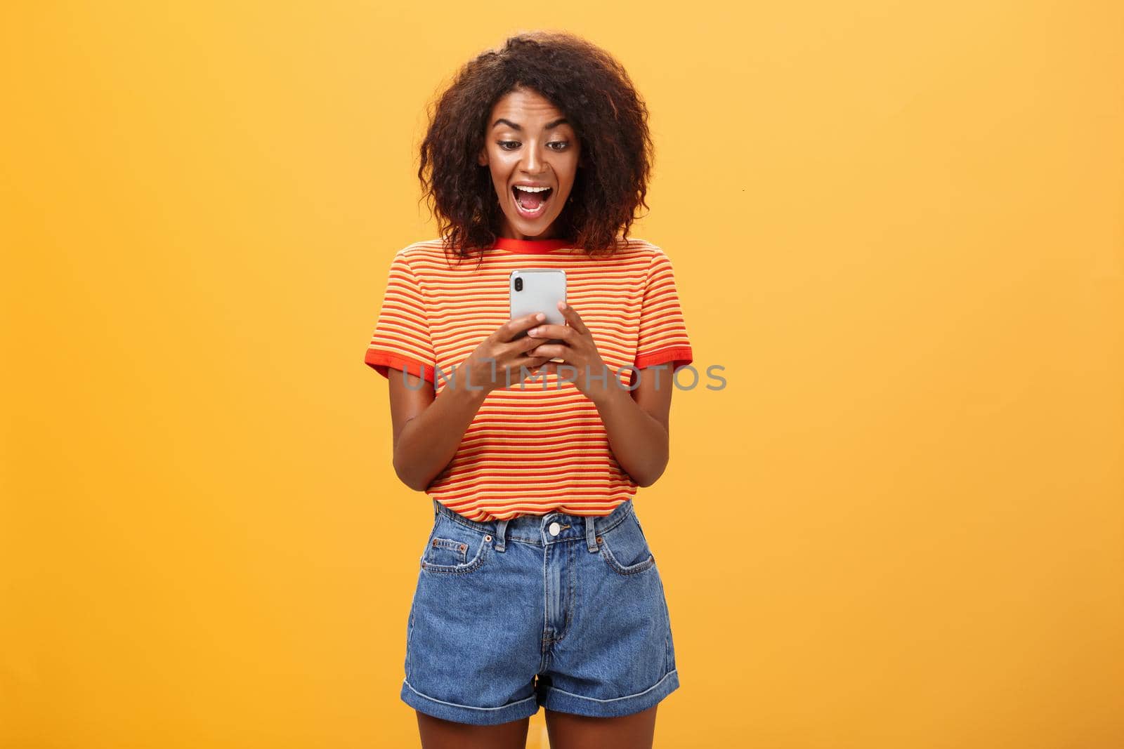 Girl expressing excitement and joy receiving awesome invitation via messages yelling from delight and happiness looking at smartphone screen impressed and thrilled over orange background.