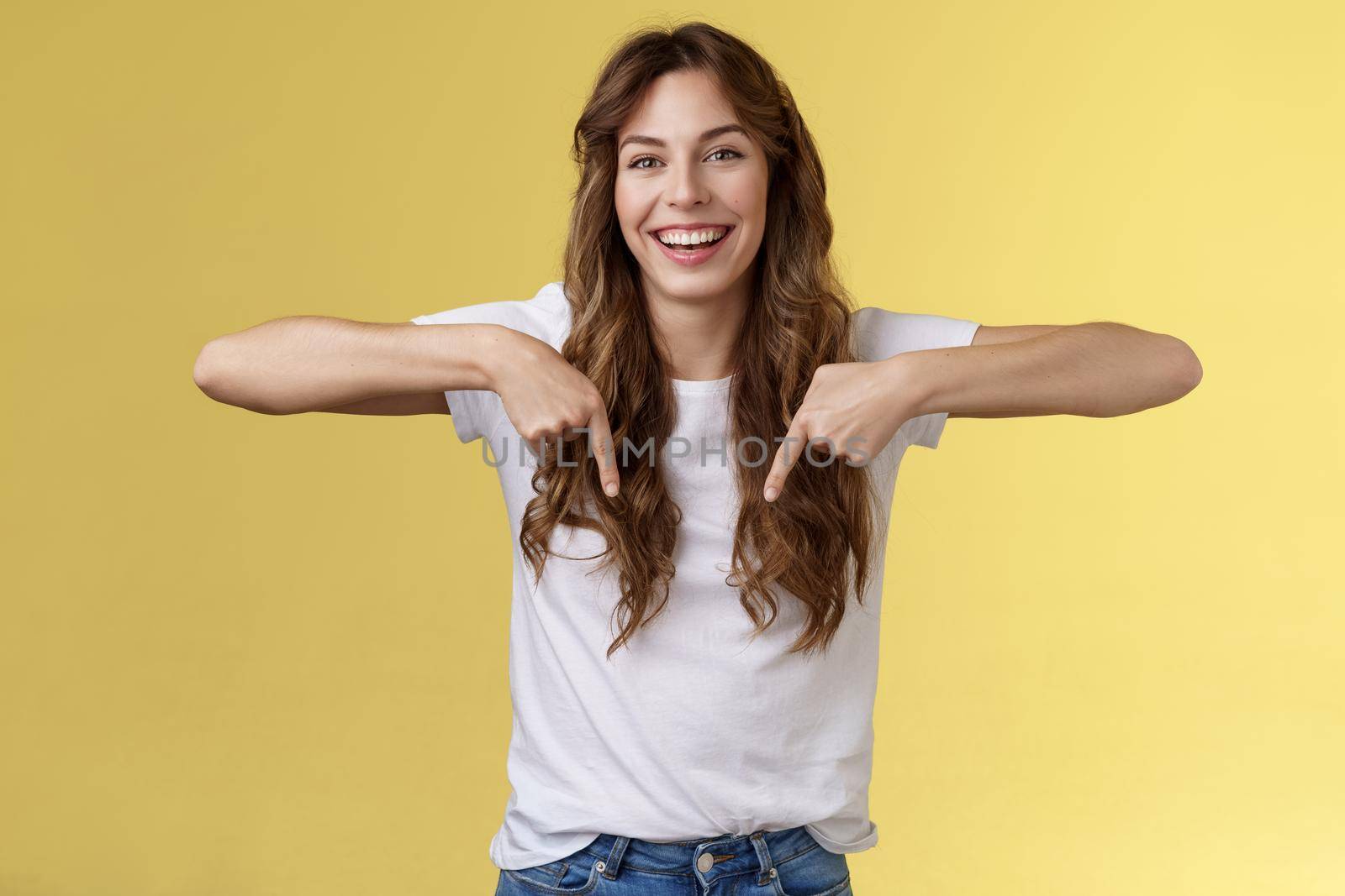 Cheerful lively happy funny girl long beautiful hair pointing down index fingers indicate bottom promo laughing joyfully share positive emotions recommend product advertising yellow background.