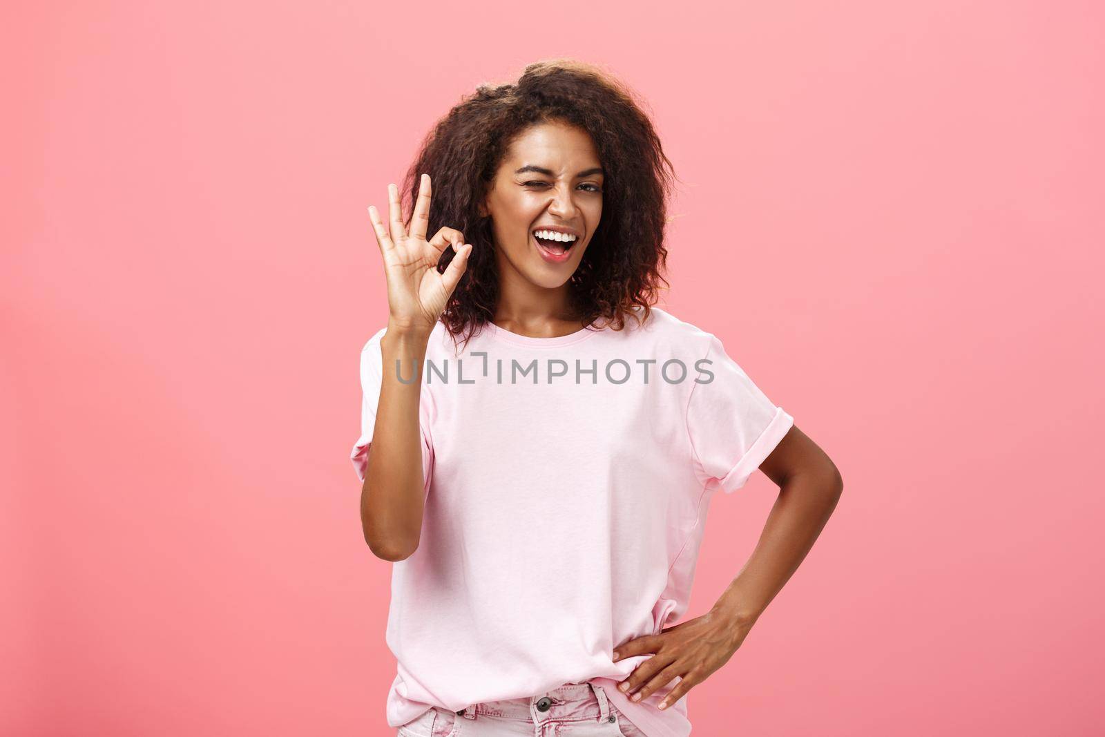 Girl got us covered. Charming charismatic and confident african american skillful woman in stylish outfit holding hand on waist showing okay gesture and winking assured at camera over pink wall by Benzoix