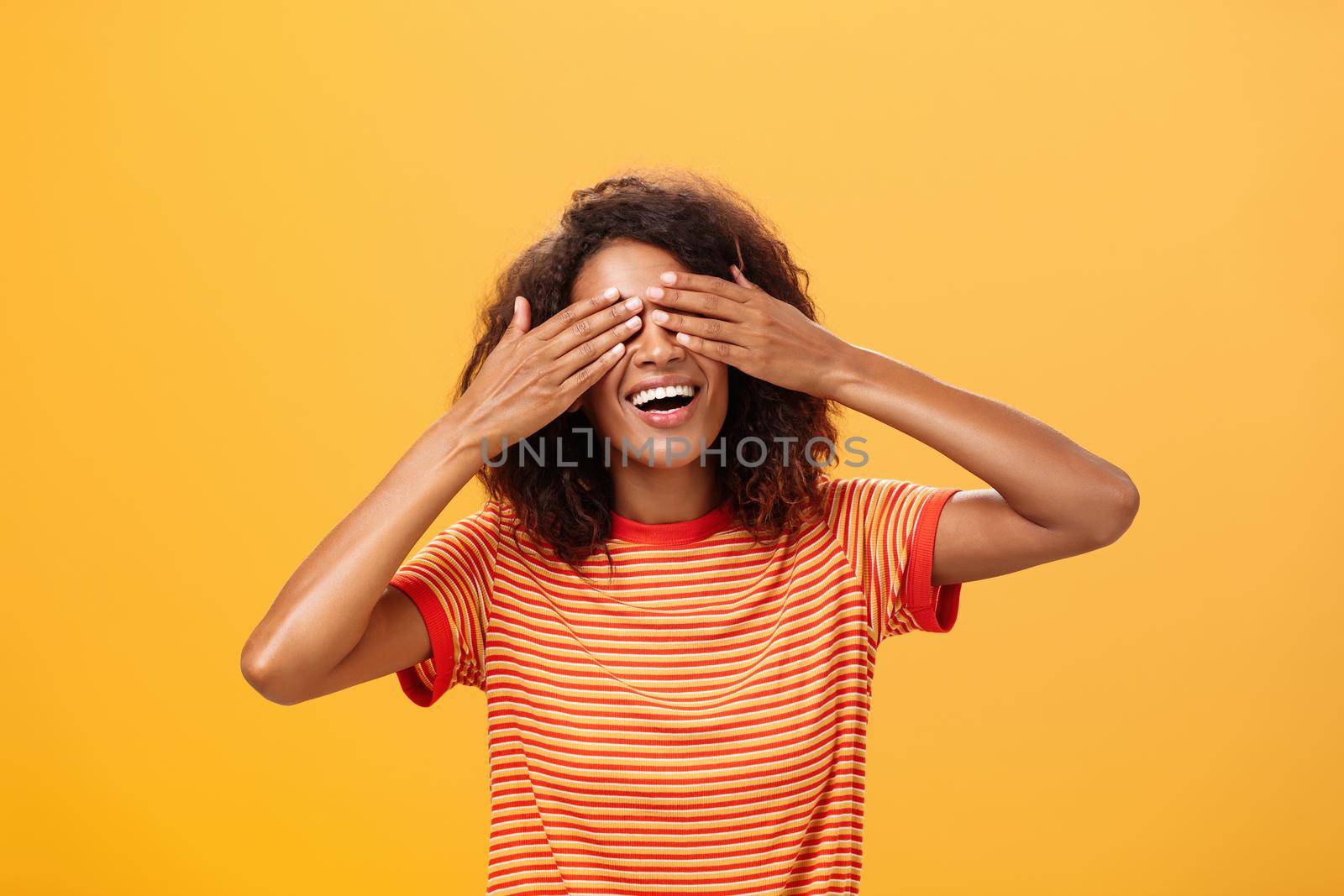 Closing my eyes and counting ten. Portrait of charming dreamy and happy funny african american curly-haired female in striped trendy t-shirt covering sight with palm and smiling waiting surprise by Benzoix