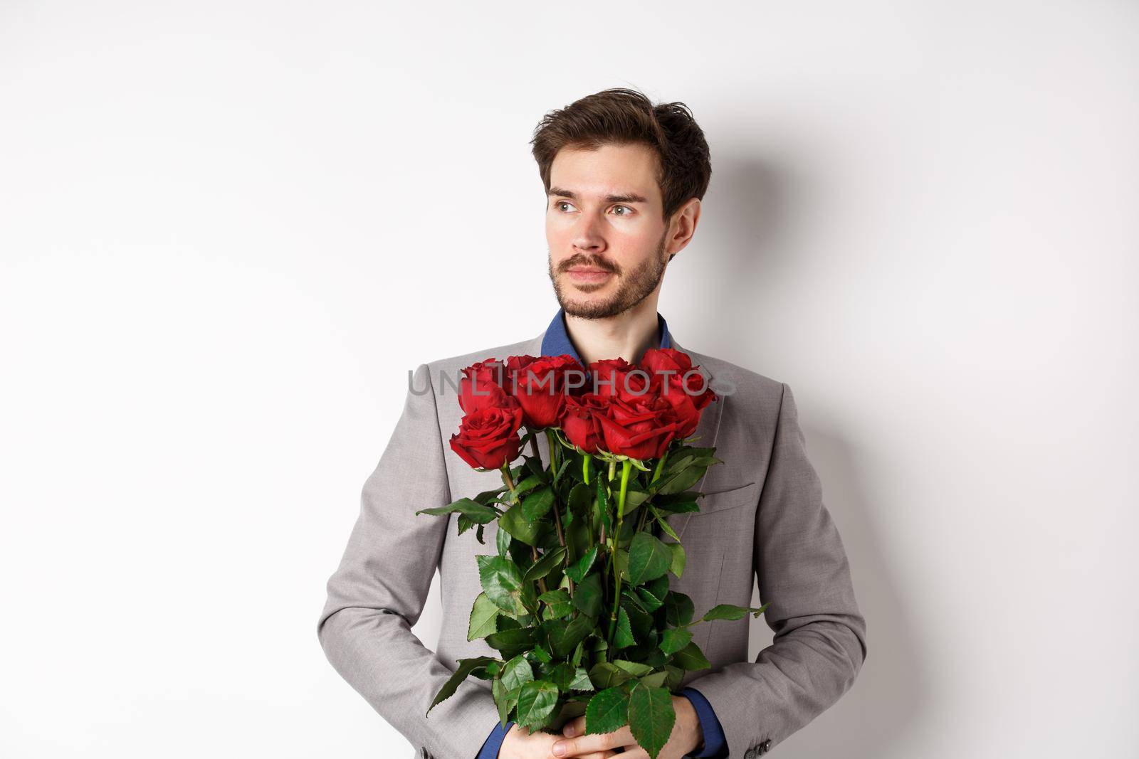 Handsome boyfriend in suit going on romantic date, holding bouquet of red roses and looking left thoughtful, standing over white background.