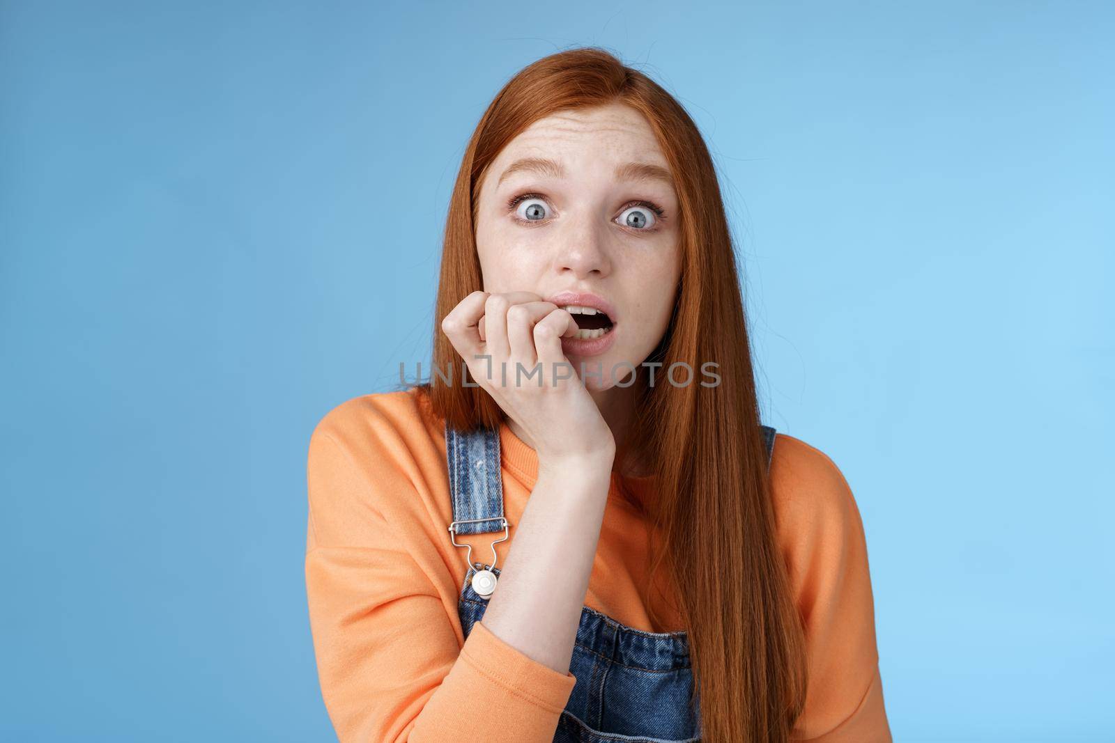 Scared unconfident anxious young trembling redhead girl wide eyes staring intense emotional biting fingernails, fan worry favorite character tv series dies standing nervously blue background.