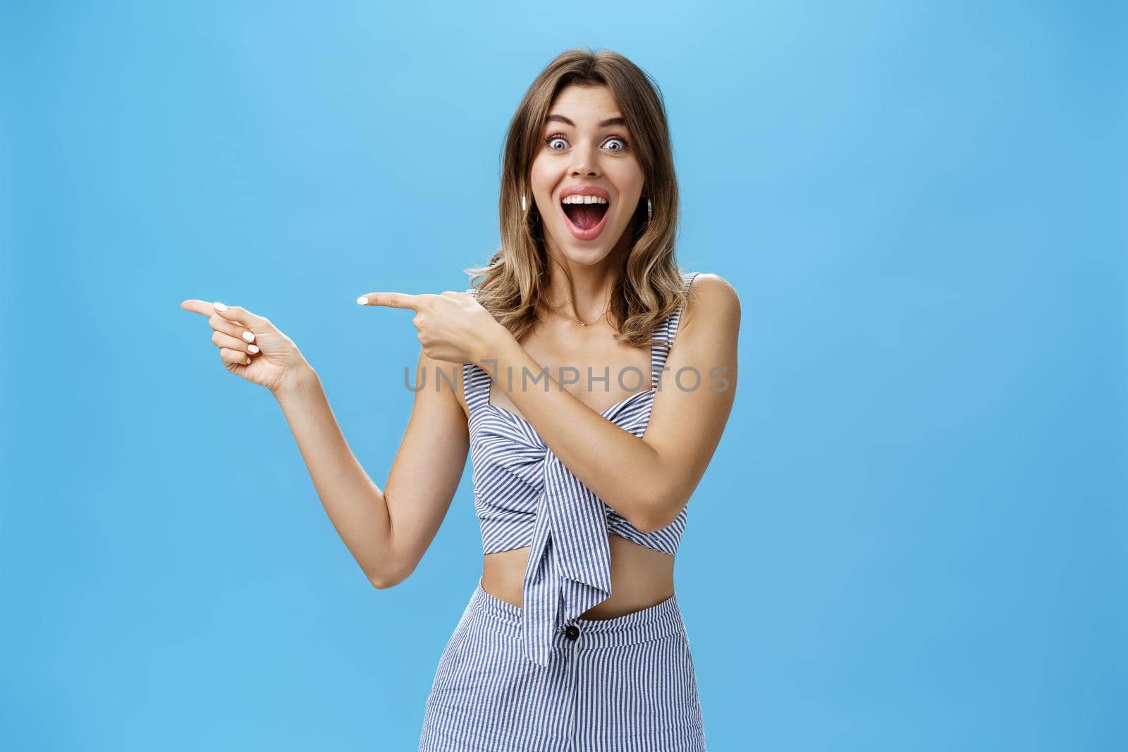 Unaltered shot of excited amused adult attractive woman with gapped teeth pointing left and smiling broadly from happiness and joy standing delighted and thrilled over blue background. Copy space
