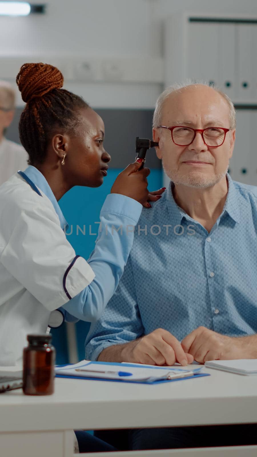 African american doctor using otoscope consulting elder man with disease in medical cabinet. Black otologist doing ear examination with professional tool on senior patient at desk