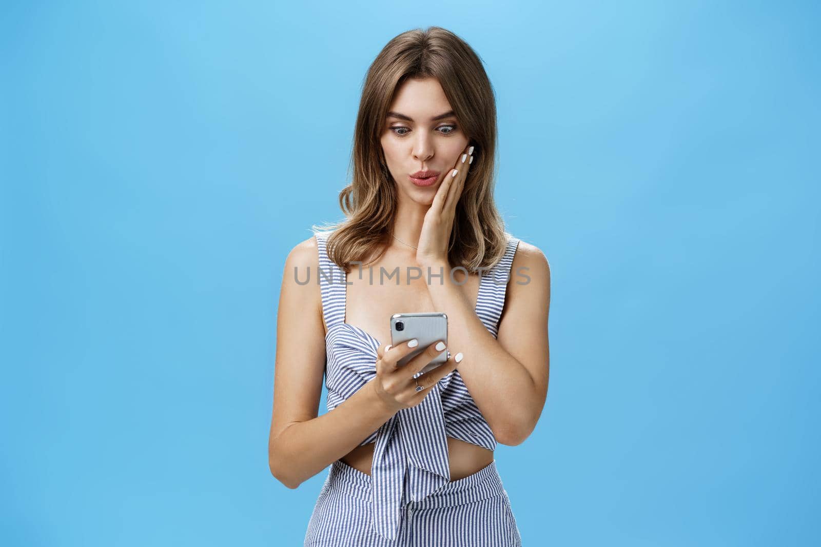 Woman learning shocking rumor receiving message in social network from friend reading news from smartphone folding lips staring excited at smartphone screen pressing palm to cheek from amazement by Benzoix