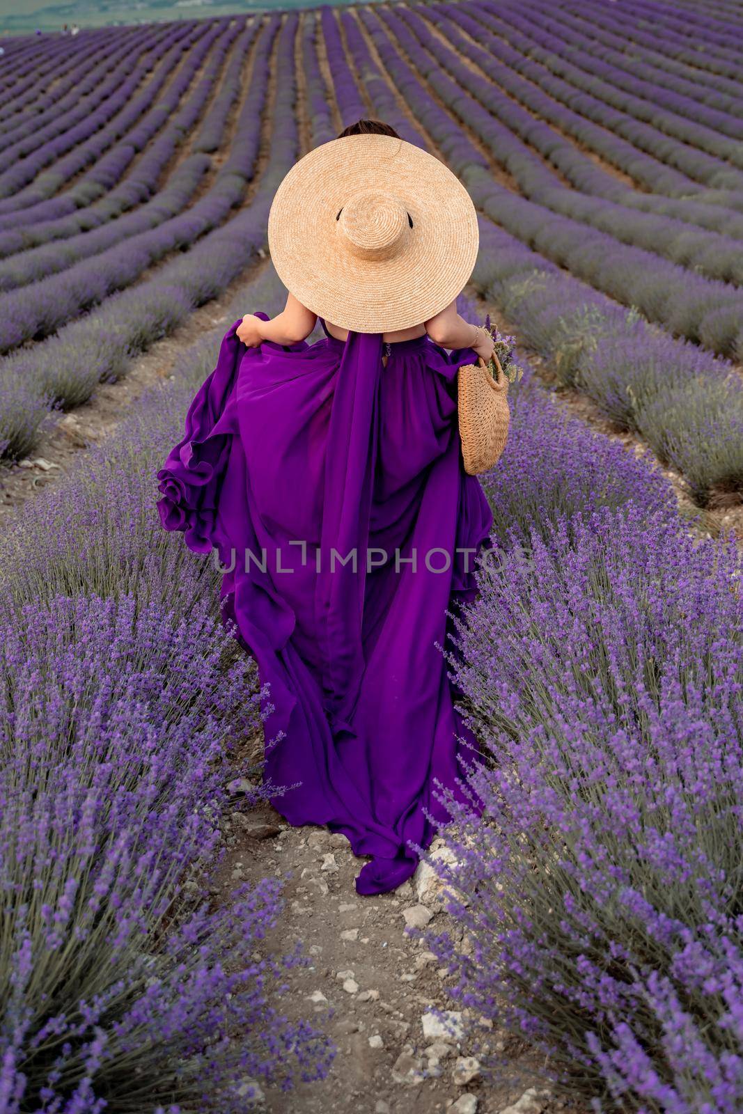 A young beautiful girl in a purple flying dress stands on a blooming lavender field. Rear view. The model has a straw hat.