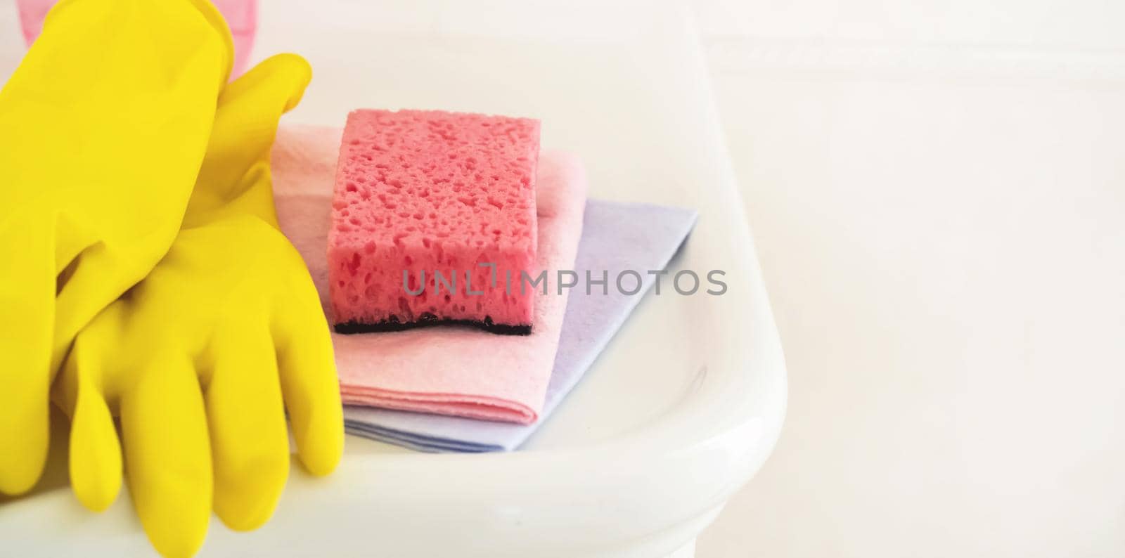 Household cleaning items, such as rubber yellow gloves, several types of rags and sponges, lie on a clean white washstand in the bathroom.